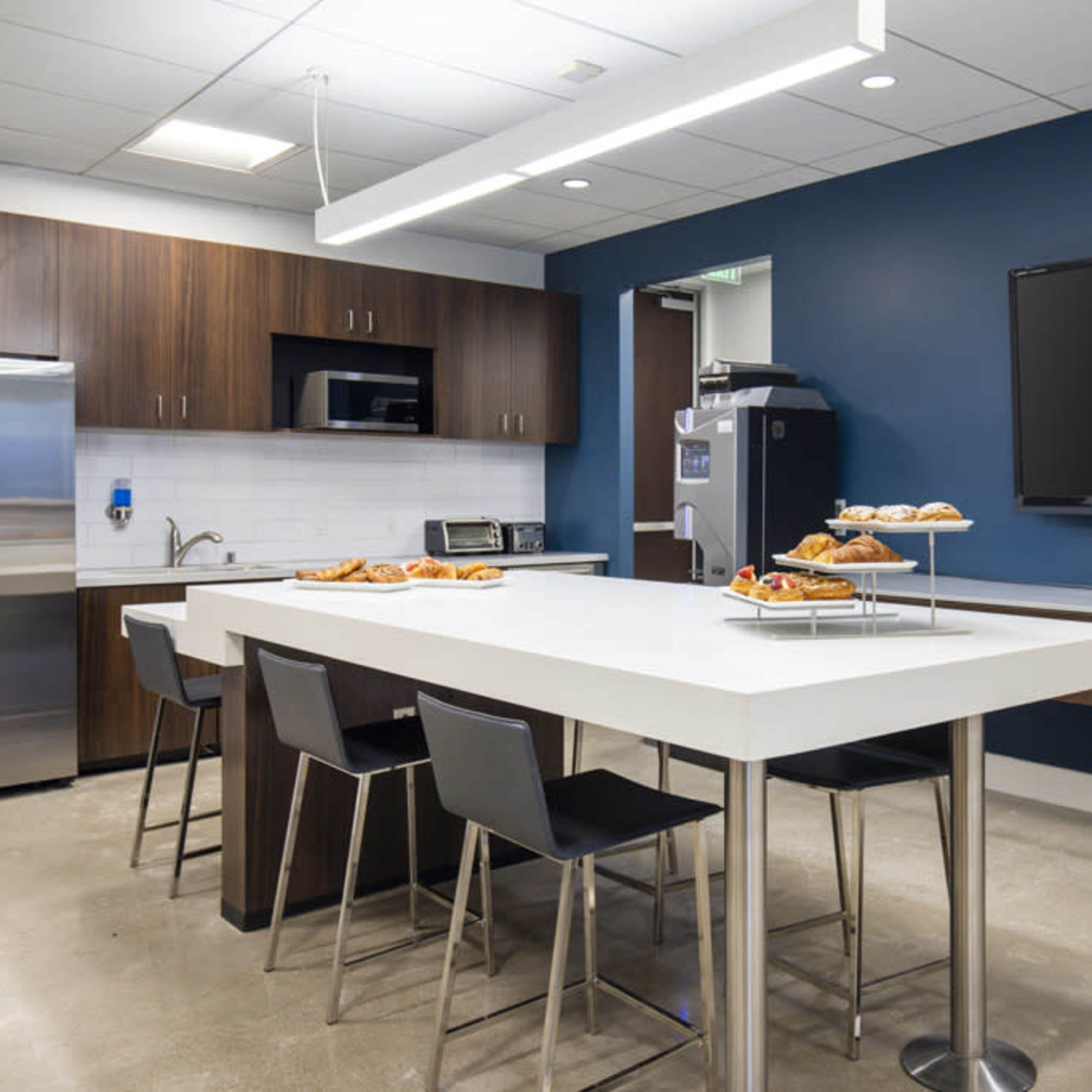 A modern kitchen area features stainless steel appliances, a central table, and a display of pastries on a tray.