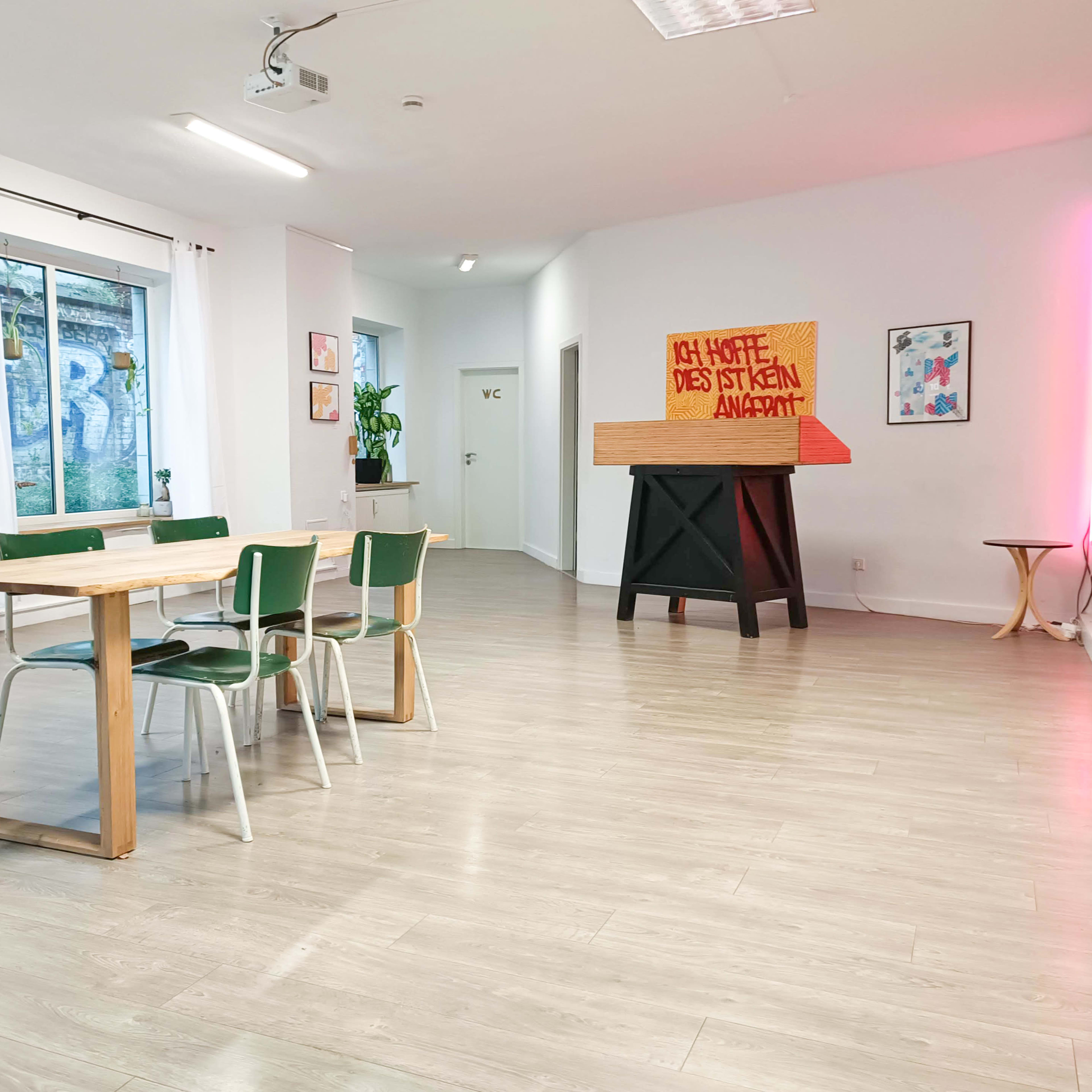 A modern, minimalist interior space with a wooden table surrounded by chairs, and a large, colorful artwork placed on an elevated stand against a white wall.