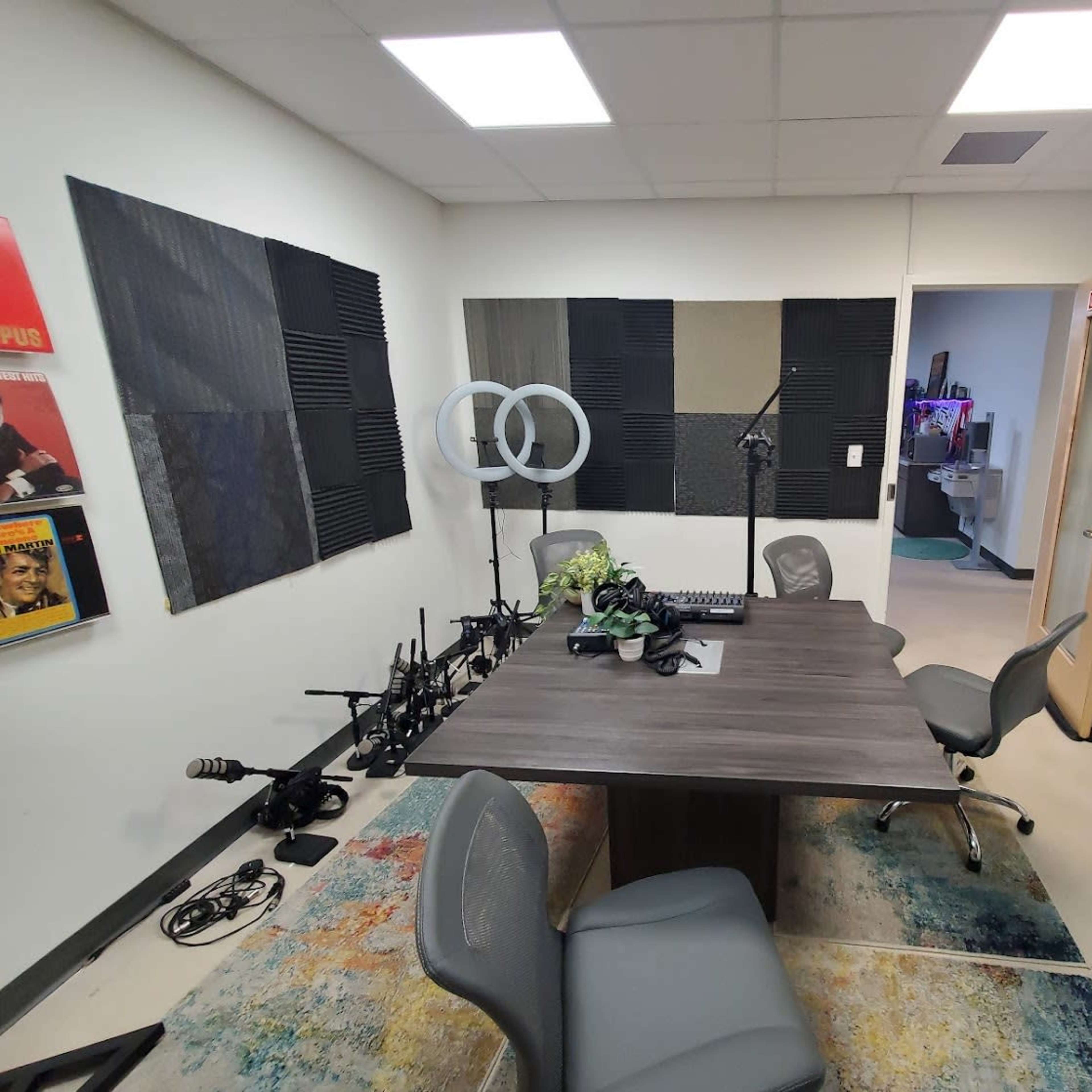 A small recording studio with a large table, two chairs, soundproofing panels on the walls, and various audio equipment arranged around the room.