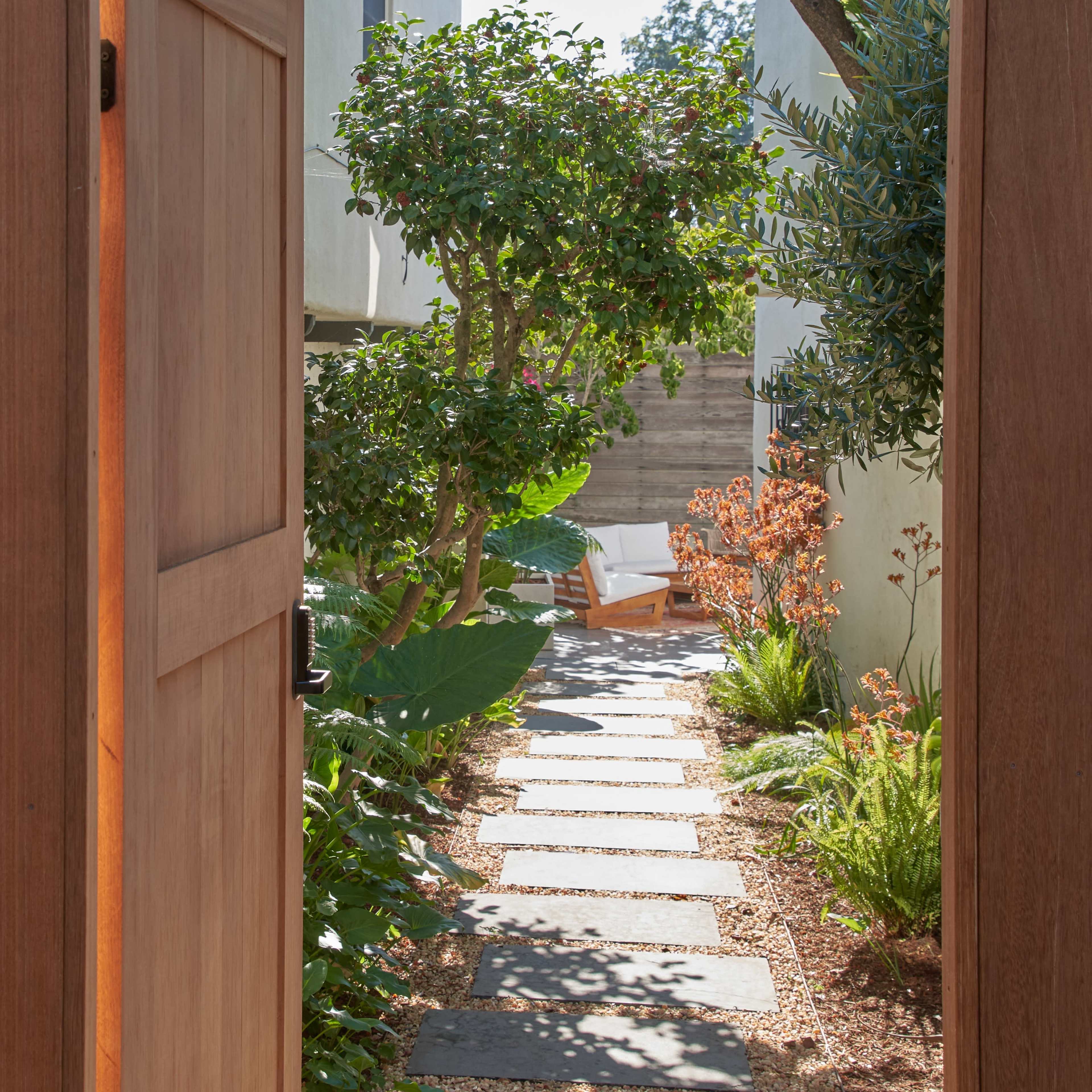 A wooden gate opens to a stone pathway lined with plants leading to a chair under a tree.
