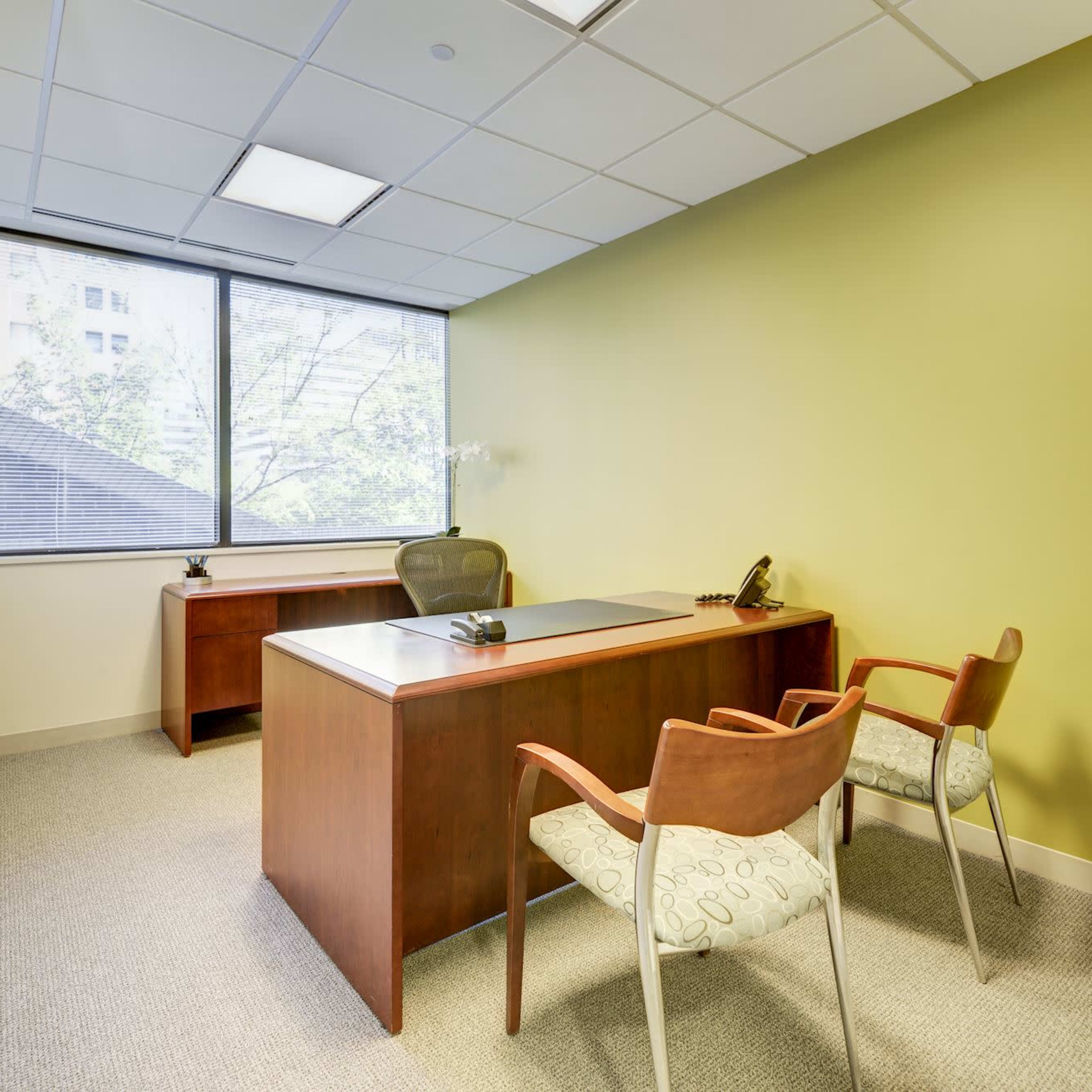 A small office with a wooden desk, an office chair, and two upholstered chairs, set against a green wall and a large window.
