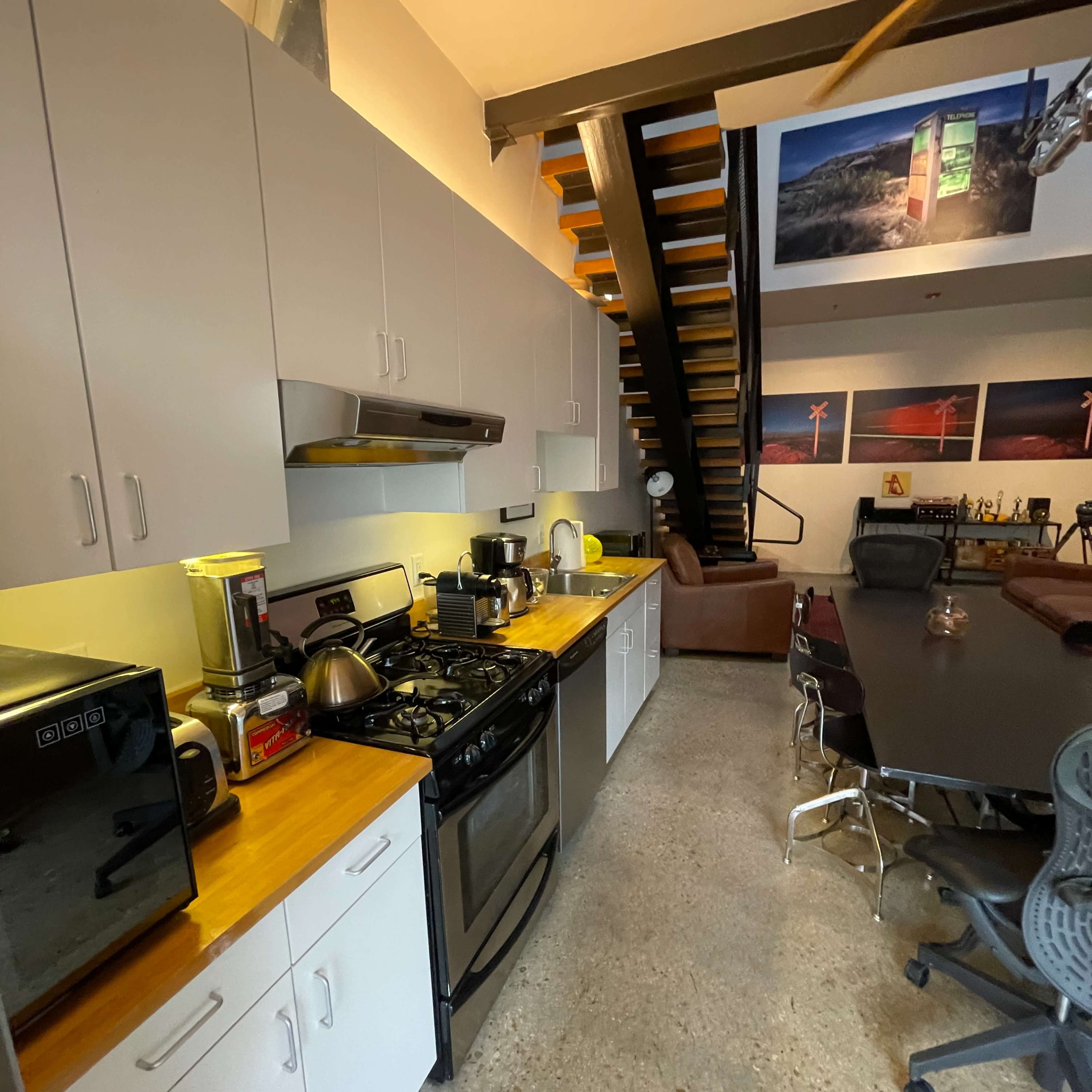 A modern kitchen with a gas stove, various appliances, and a long wooden countertop, adjacent to a dining area featuring a large table and a staircase in the background.