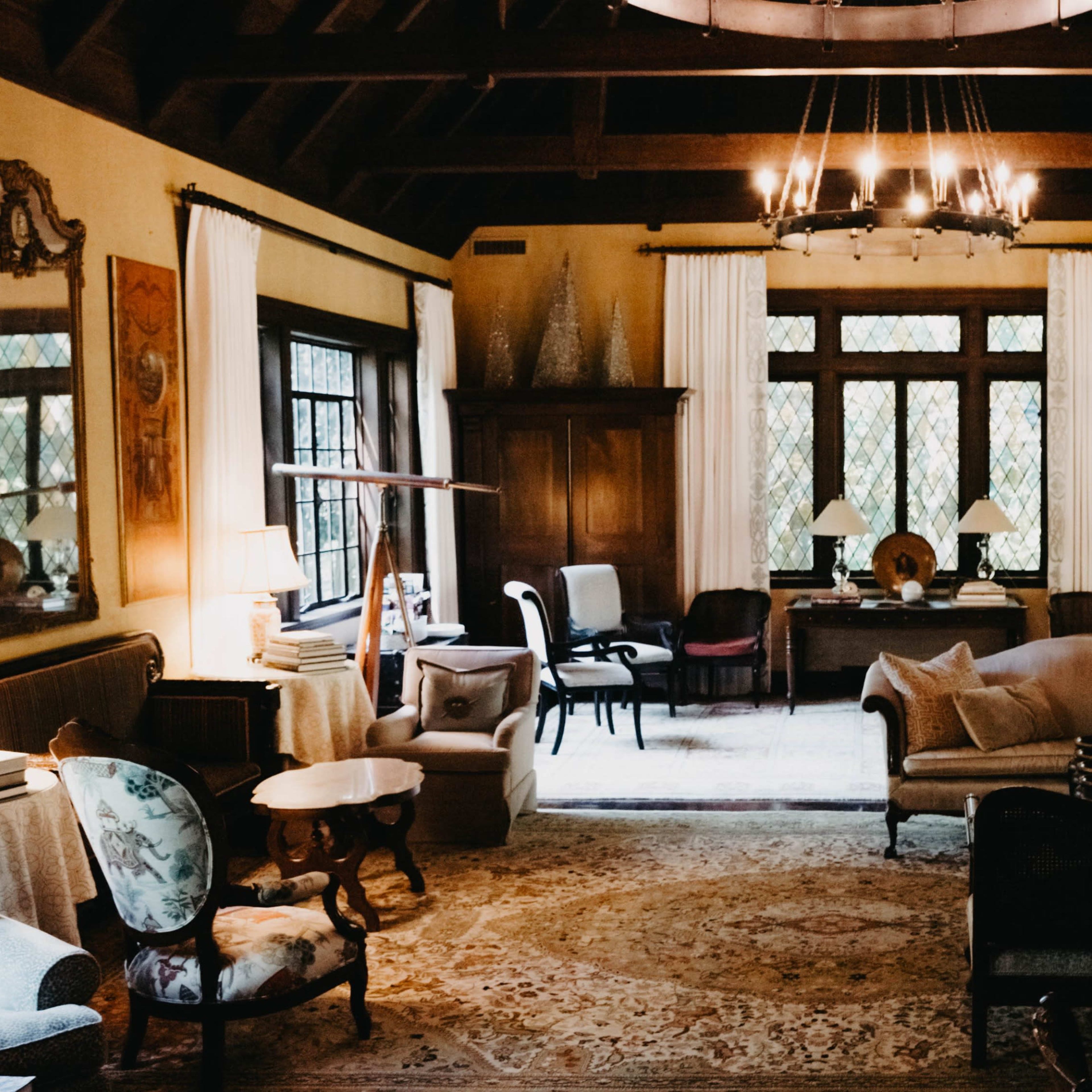 A spacious living room with high wooden ceilings, large windows, and a mix of antique furniture arranged around a central area rug.
