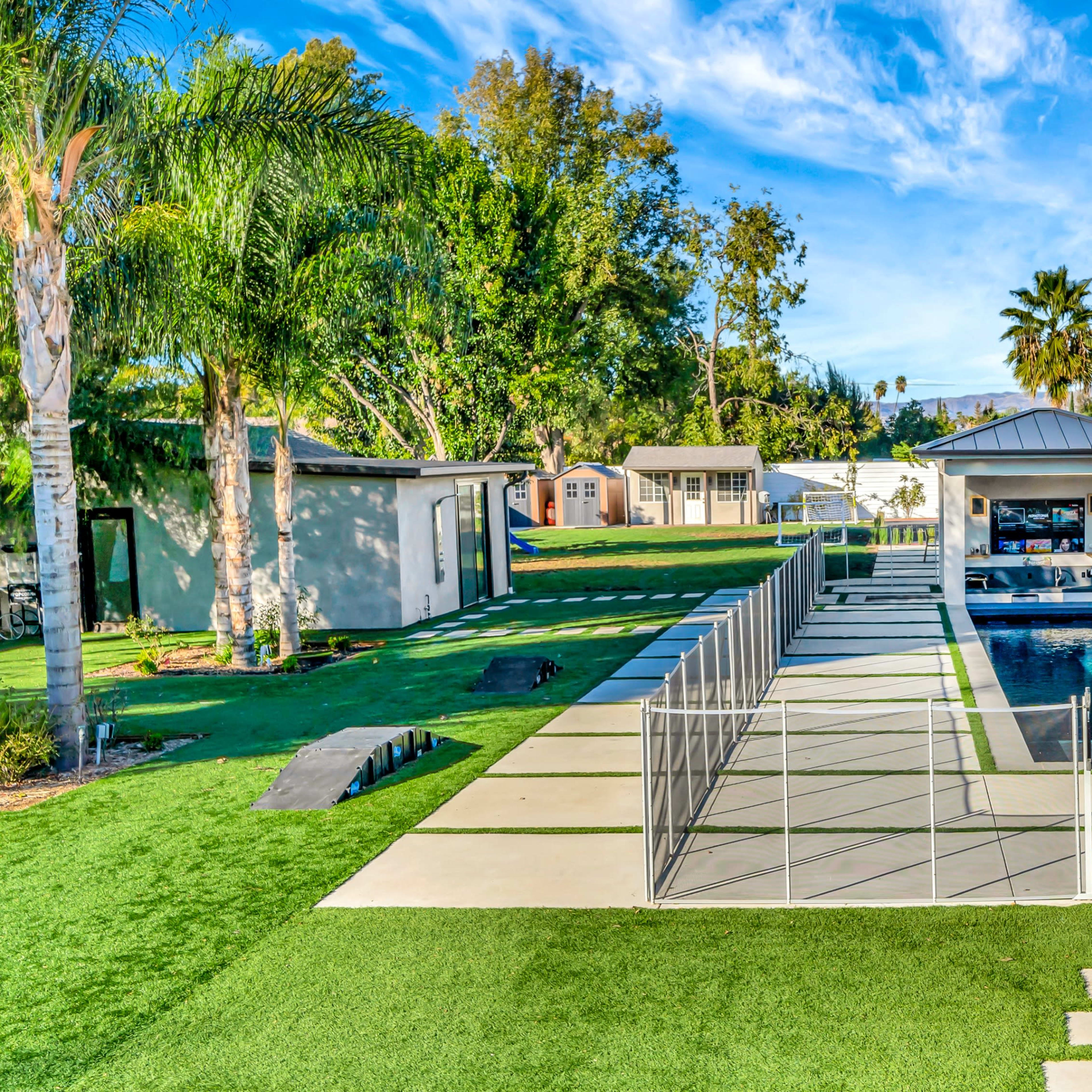 A modern backyard with a swimming pool, surrounded by palm trees, green grass, and a poolside area featuring lounge chairs and a gazebo.