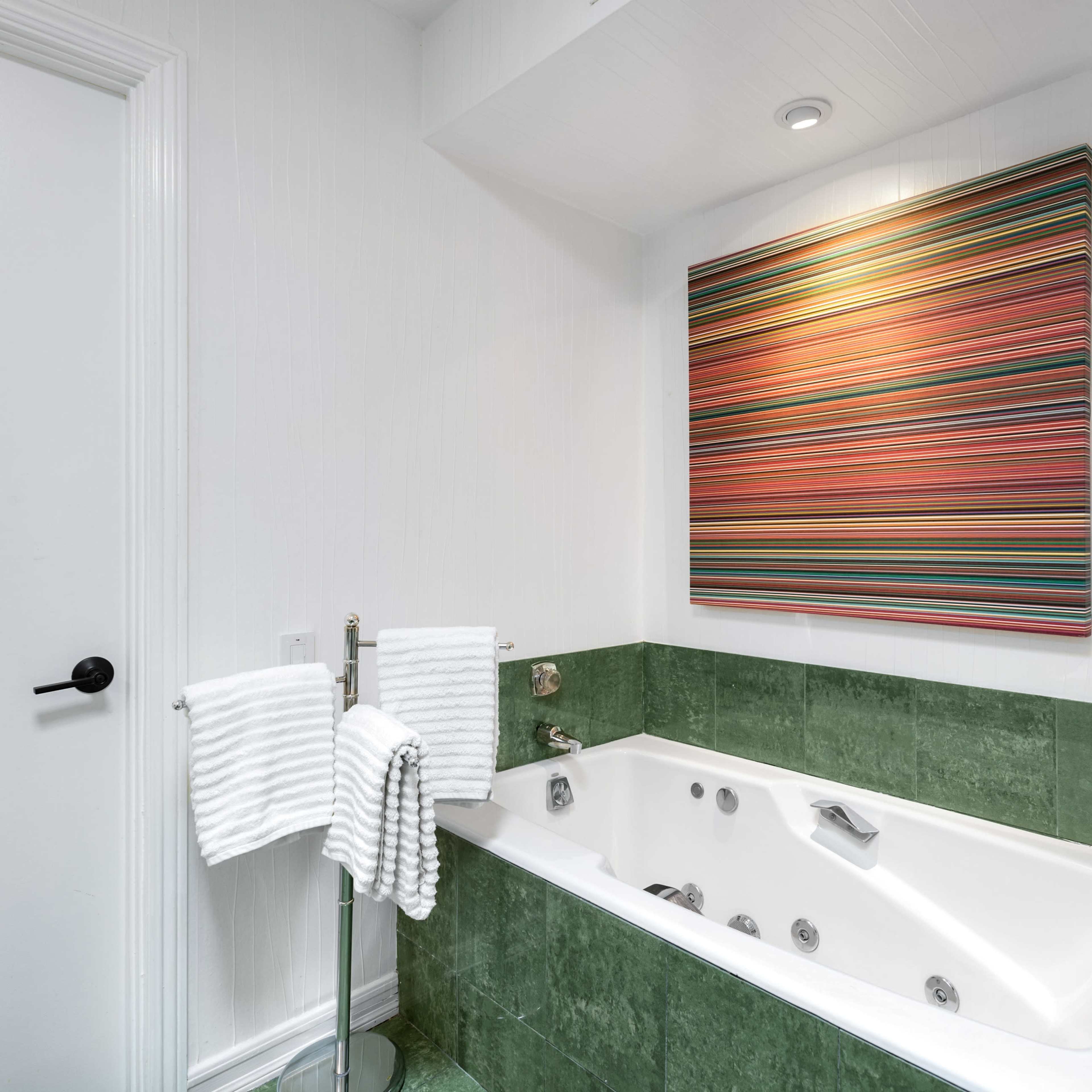 A bathroom with a green-tiled jacuzzi tub, two white towels hanging on a rack, and a colorful striped artwork on the wall.