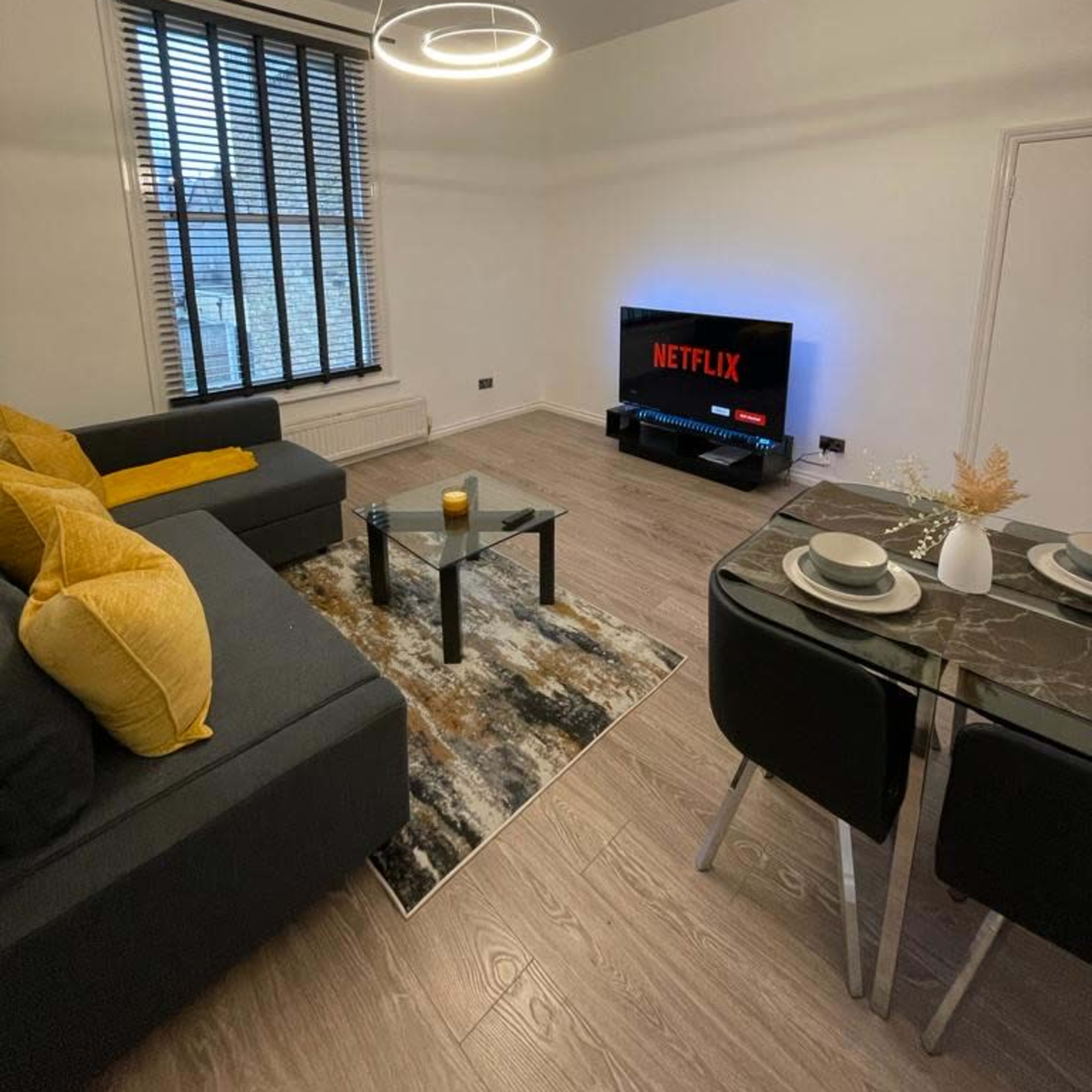 A modern living room featuring a gray sofa with yellow cushions, a round dining table set for two, and a television displaying the Netflix logo.