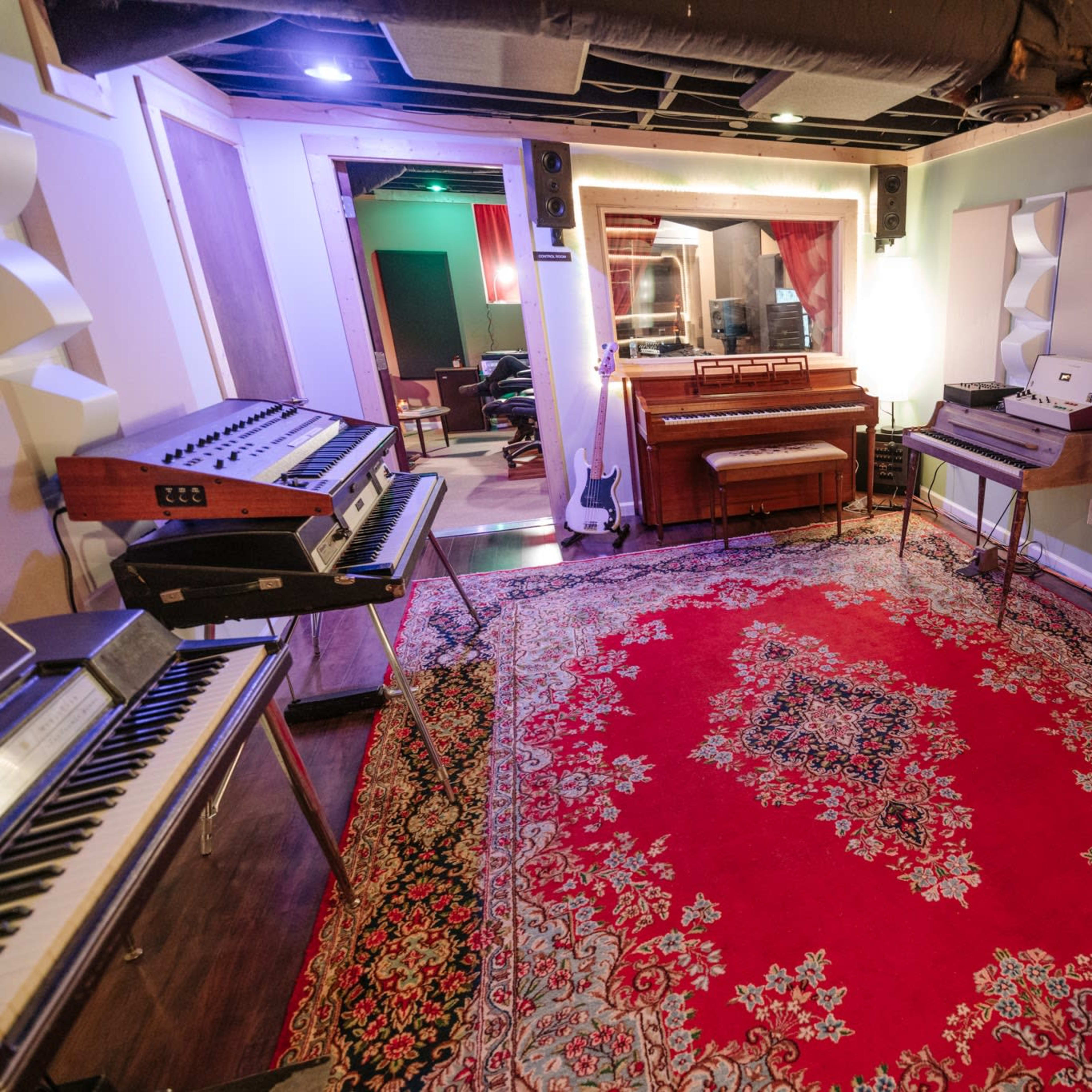 A music studio with various keyboards and synthesizers arranged around a red area rug, featuring a mirror and a recording area in the background.