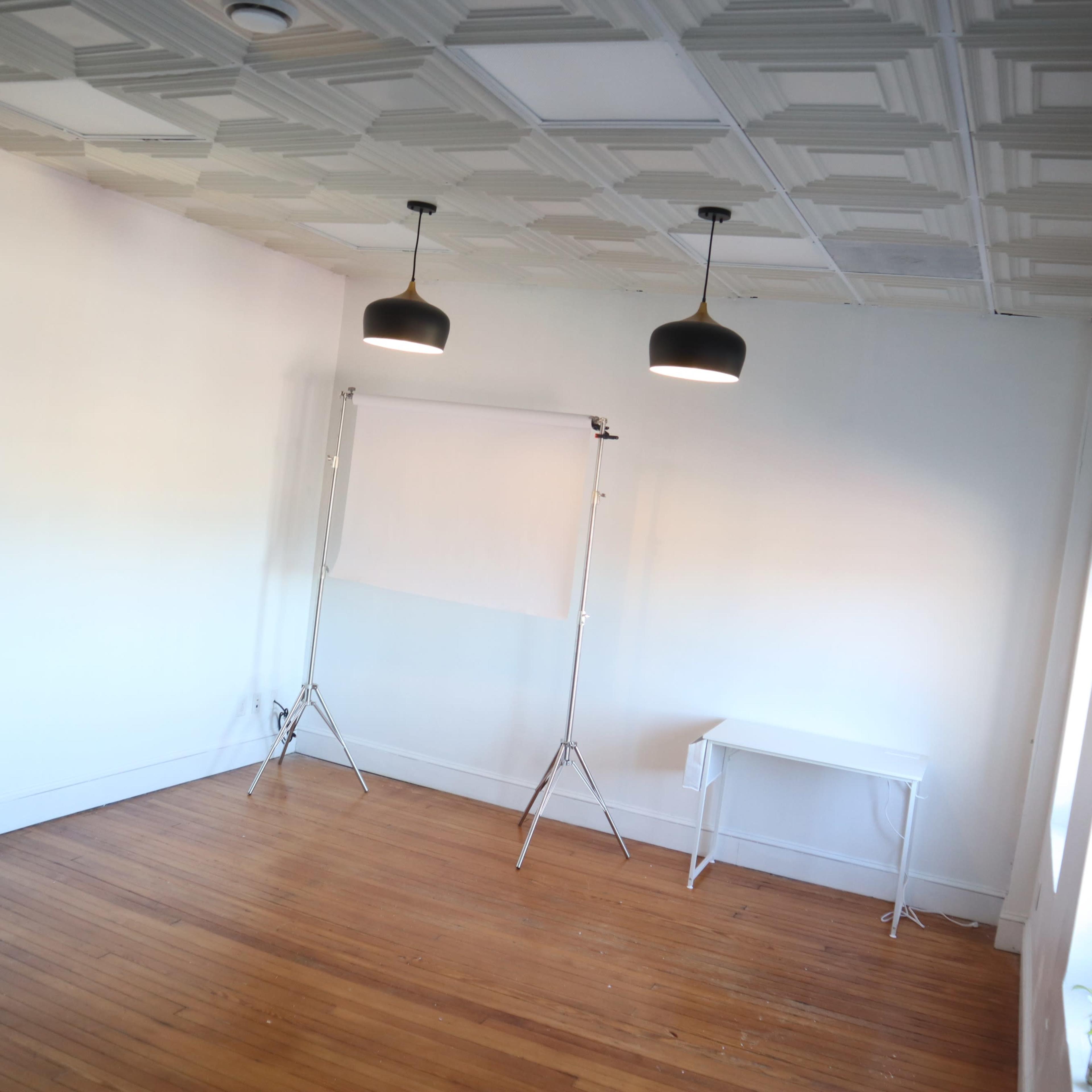 A spacious, empty room with wooden flooring, two pendant lights hanging from the ceiling, and a white backdrop stand near a small white table.