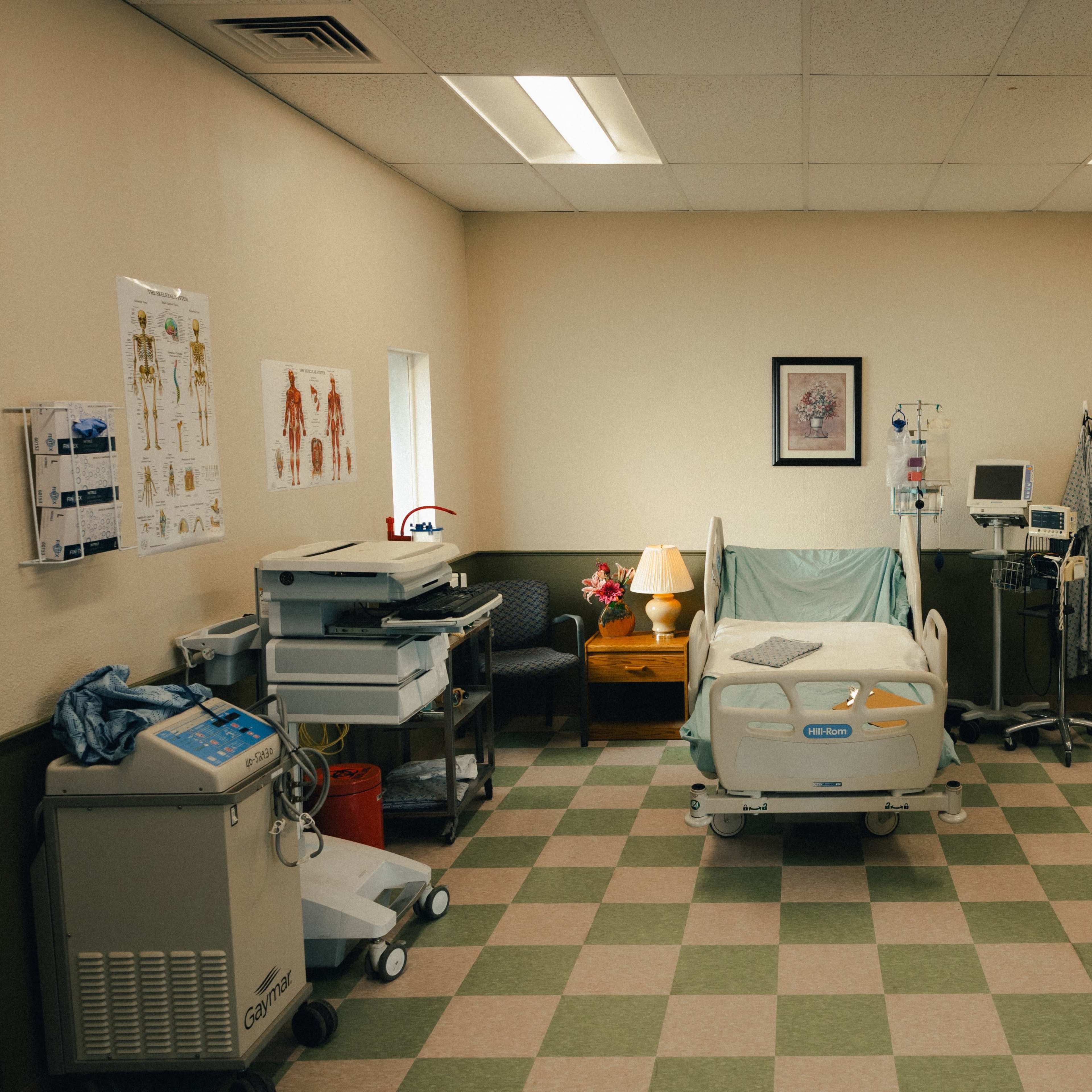 A sterile hospital room with a bed, medical equipment, a lamp on a small table, and charts on the walls.