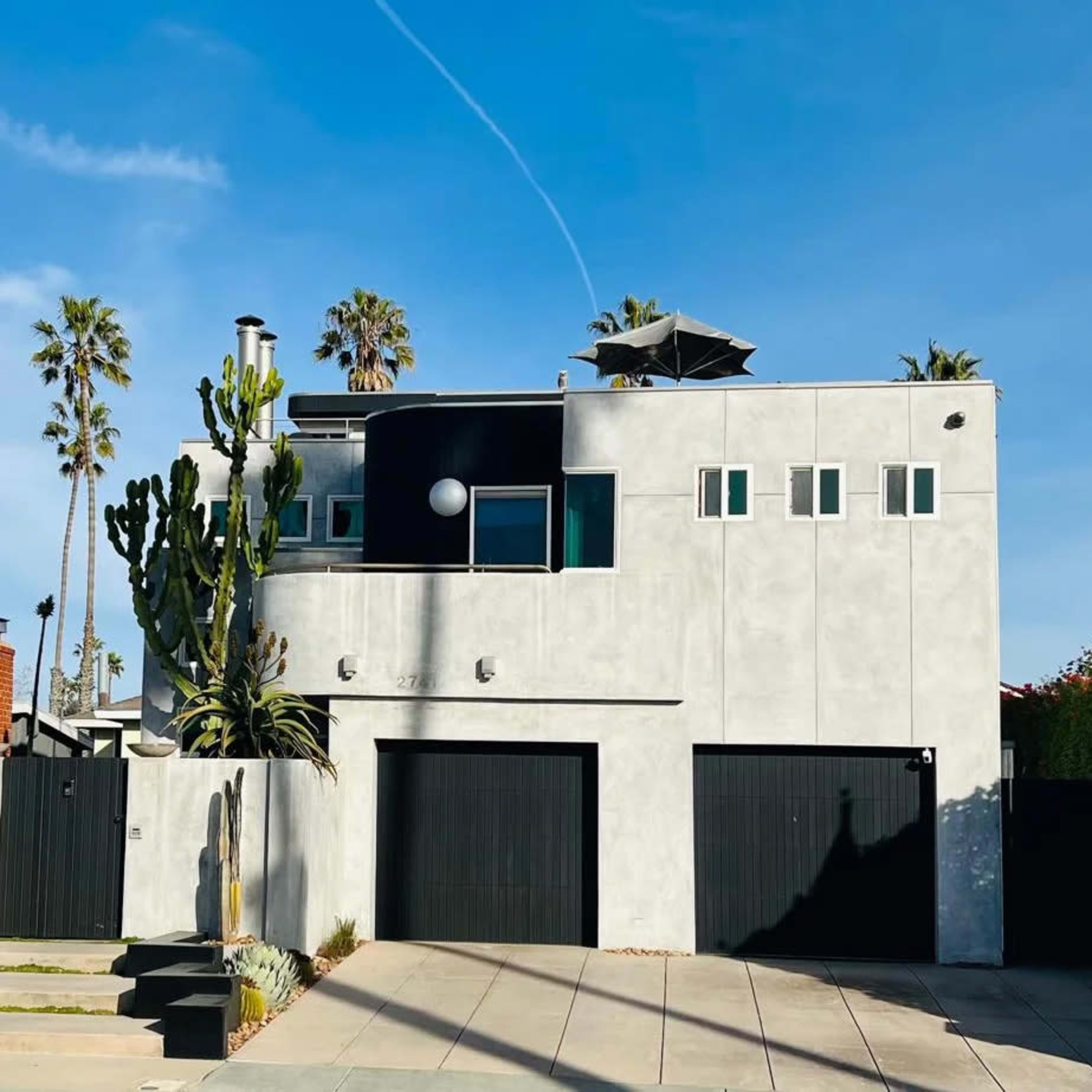A modern two-story house with a concrete exterior, featuring large windows, a flat roof, and two black garage doors, surrounded by palm trees.
