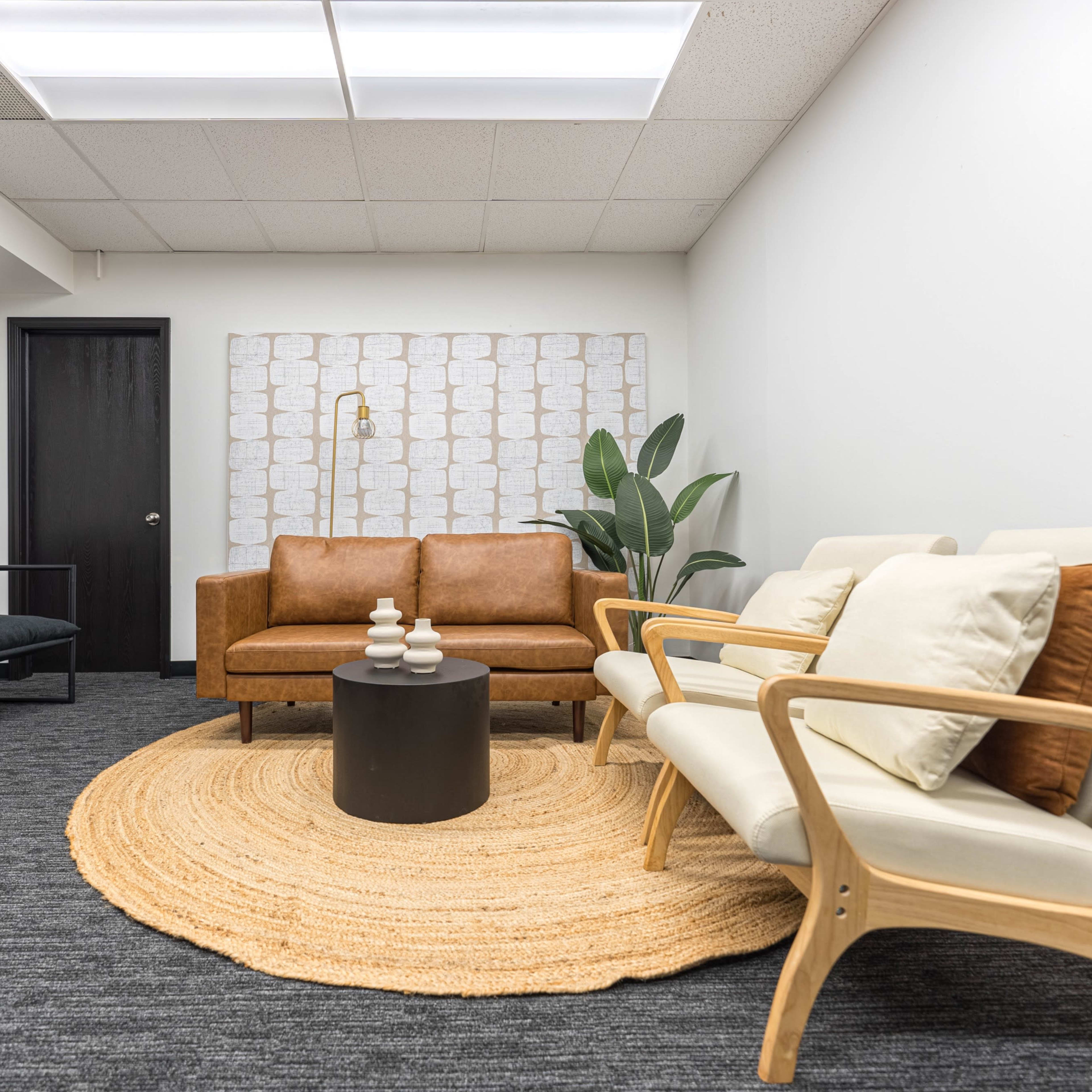 A modern waiting area with a tan leather sofa, light-colored armchairs, a round black table, and a large circular rug.