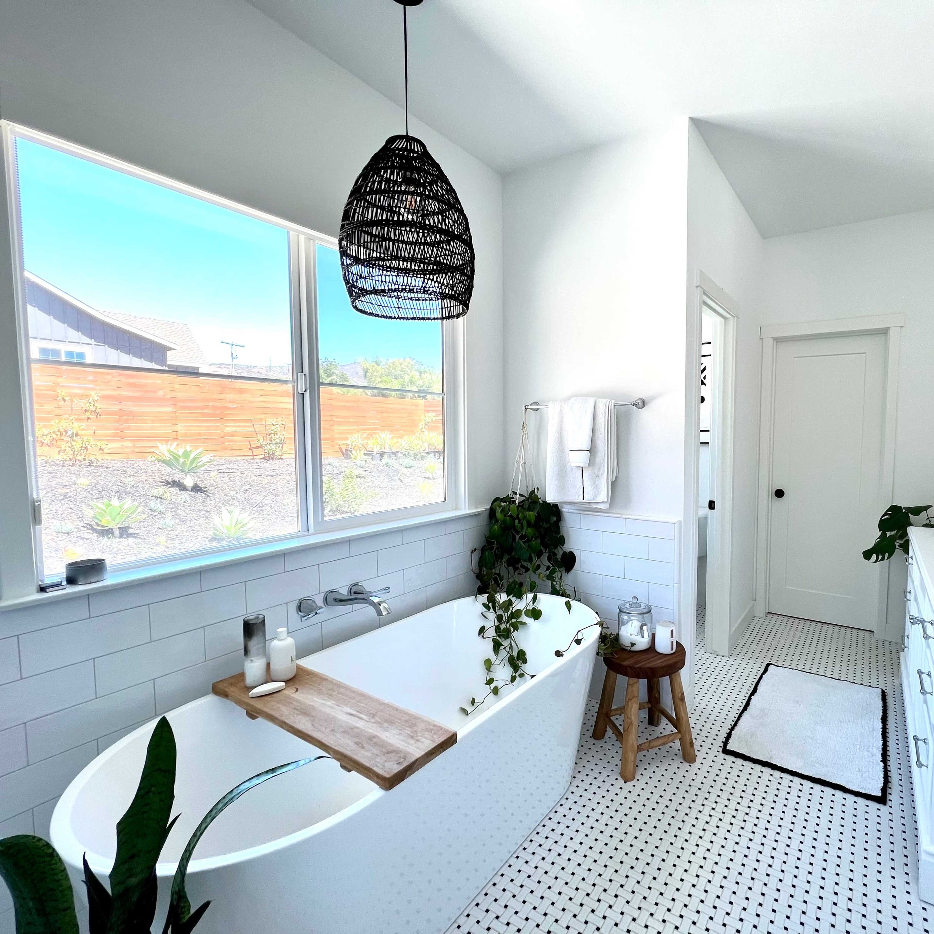 A modern bathroom with a freestanding bathtub, a large mirror, and a white vanity, featuring natural light from a window and greenery accents.