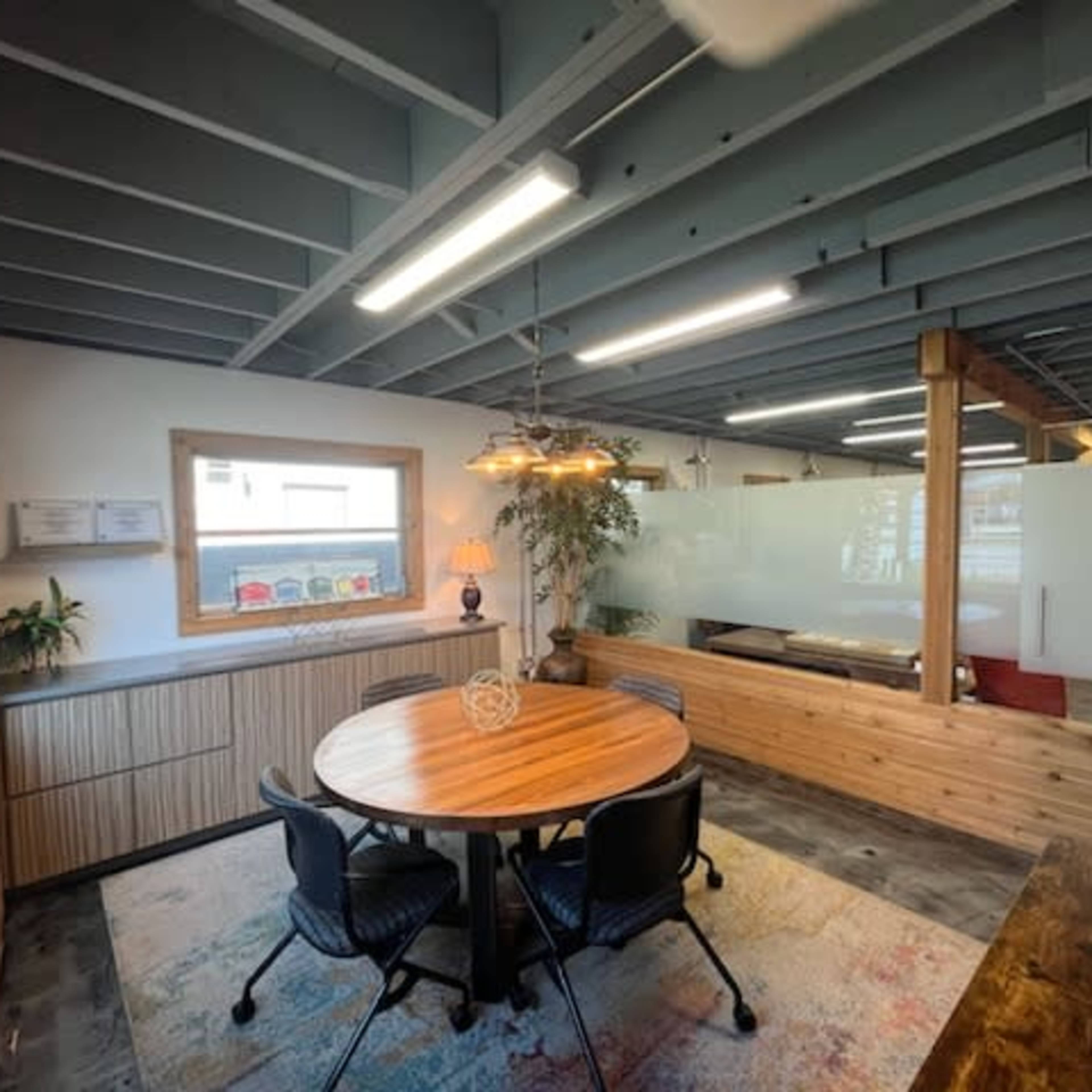 A round wooden table is surrounded by black chairs in a modern office space featuring exposed beams and large windows.