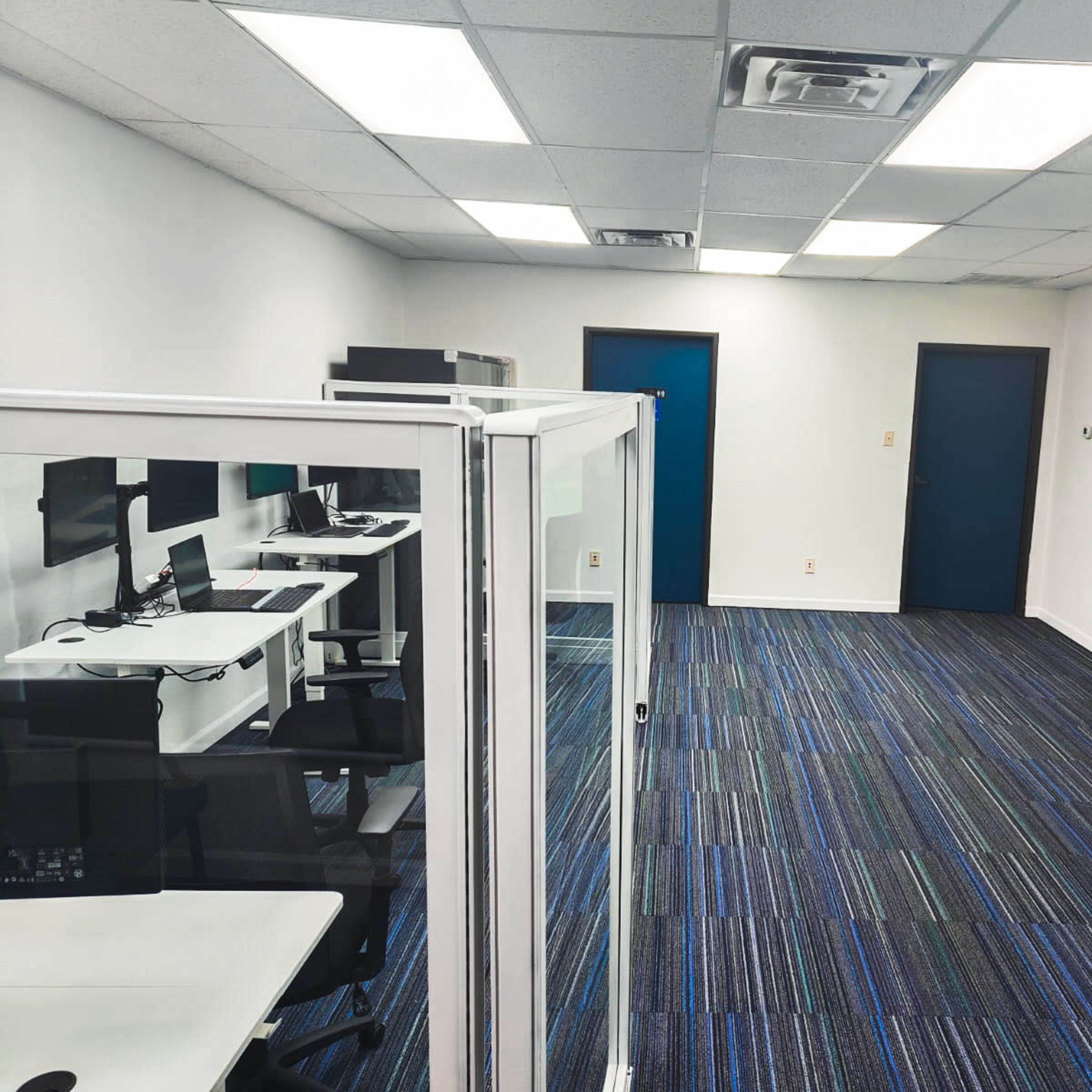 A modern office space with cubicles, desks, and blue carpet, featuring white walls and dark blue doors.