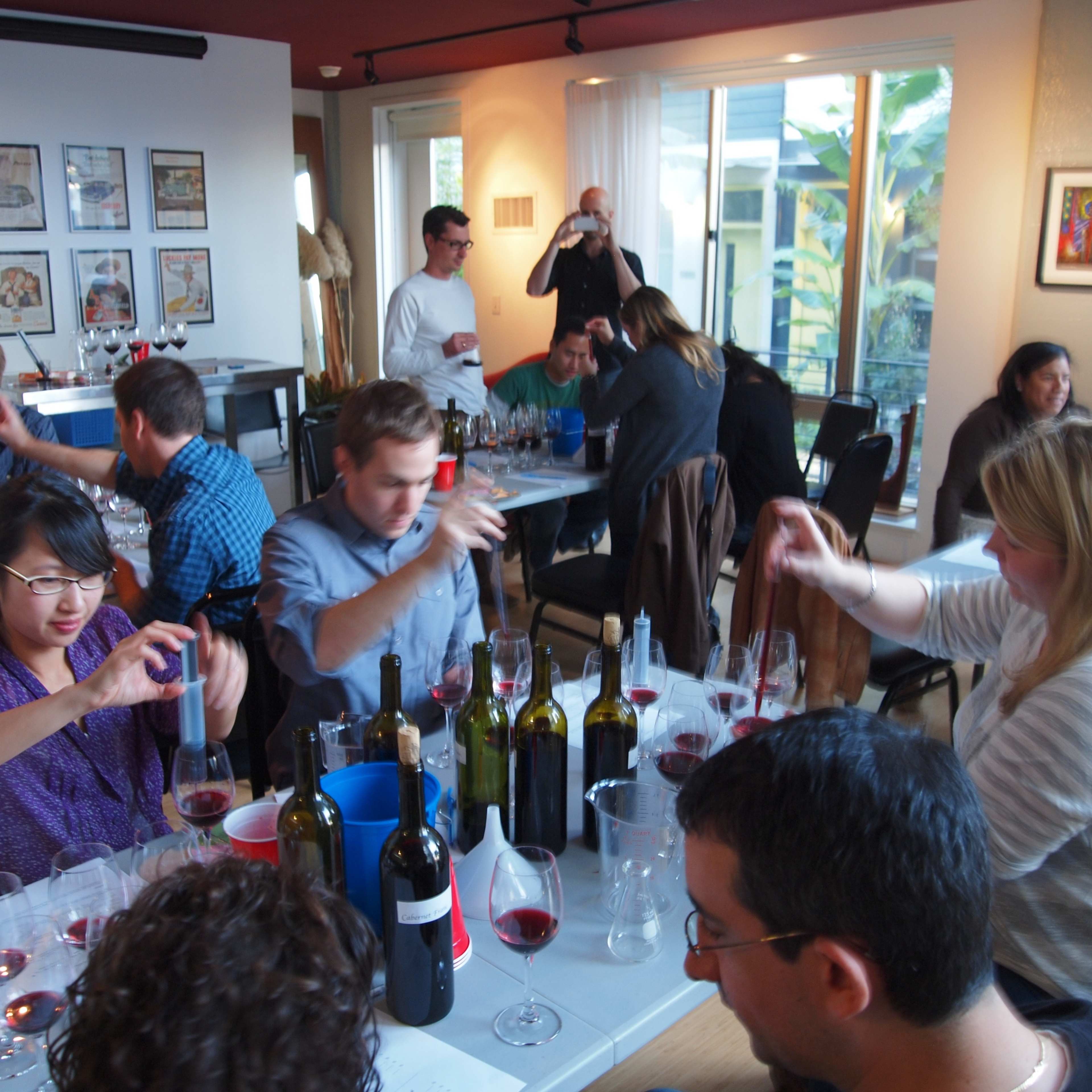 A group of people is participating in a wine tasting event around a table filled with glasses and wine bottles in a well-lit room.