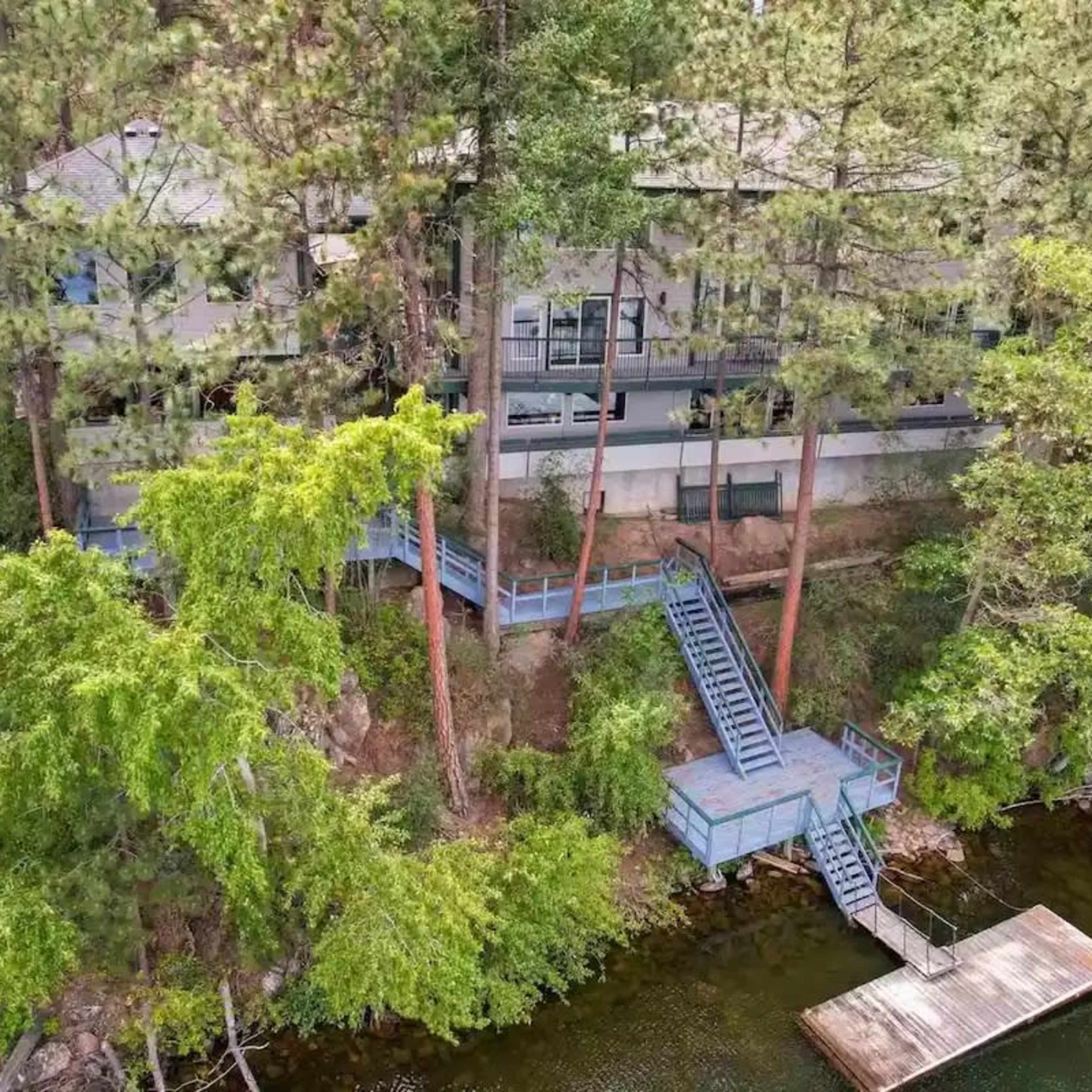 A house built on a hillside near a body of water, surrounded by tall trees, features a staircase leading down to a dock.