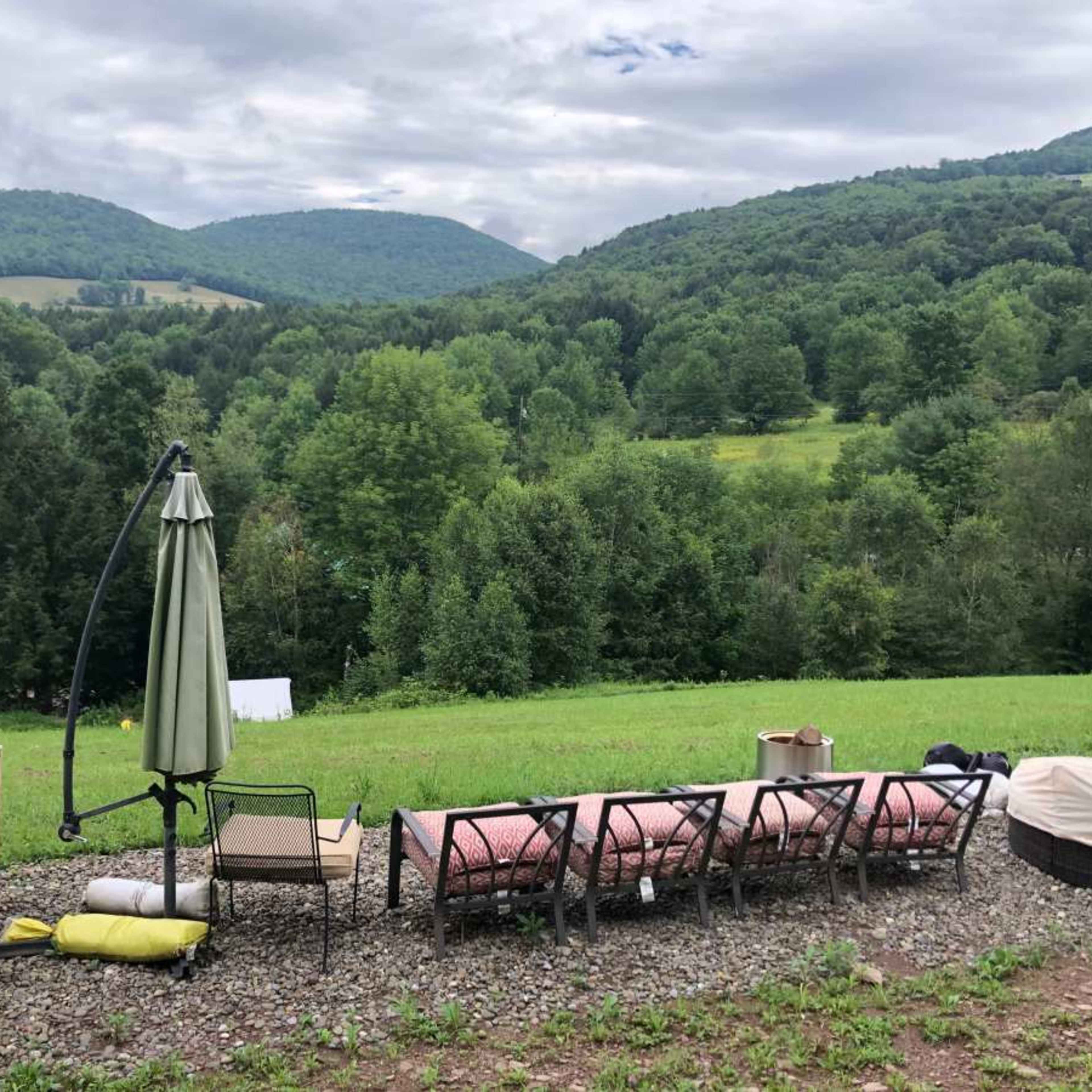 A row of outdoor chairs and a table overlook a lush, green valley surrounded by mountains under a cloudy sky.