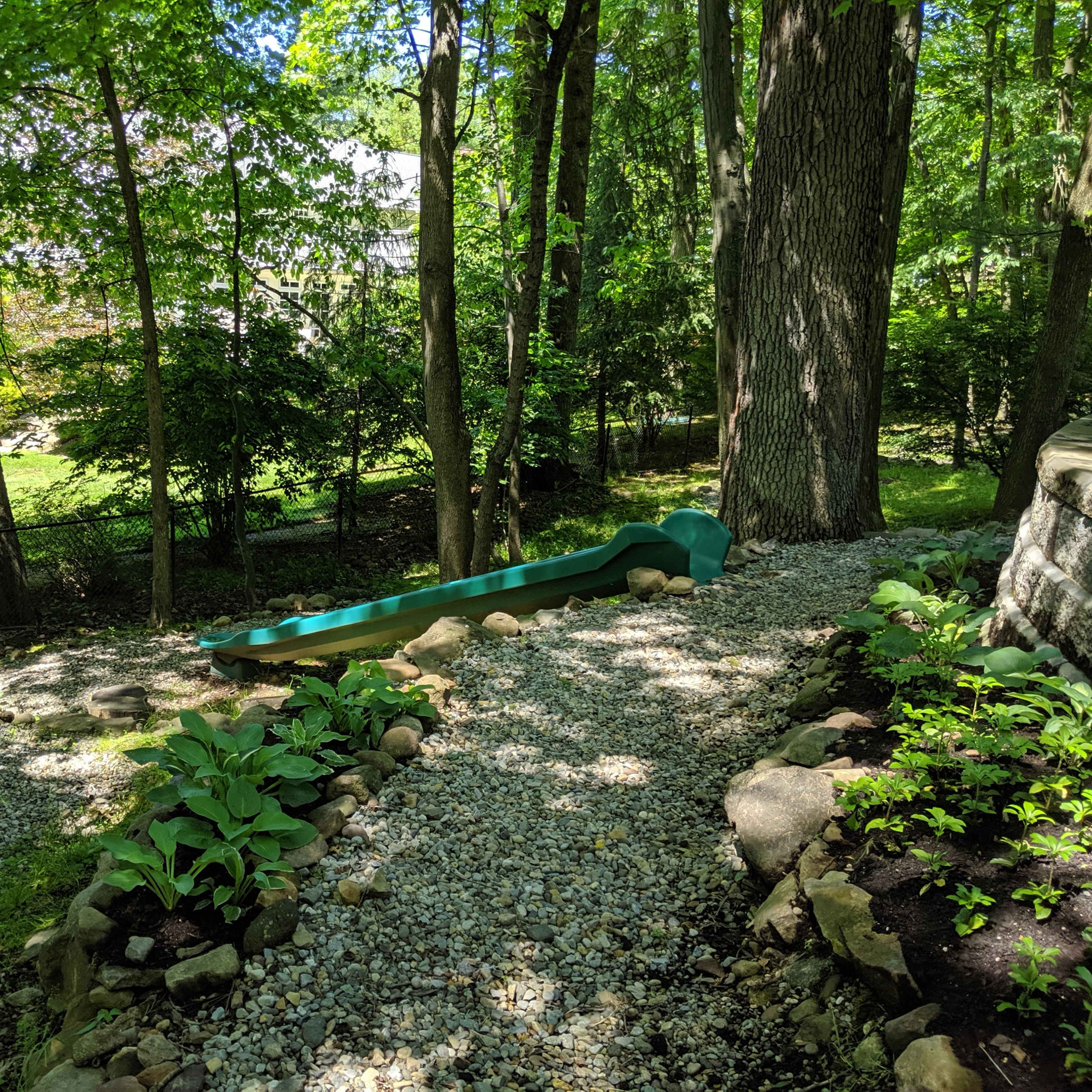 A gravel pathway winding through a landscaped garden with a slide and a trampoline among the trees.