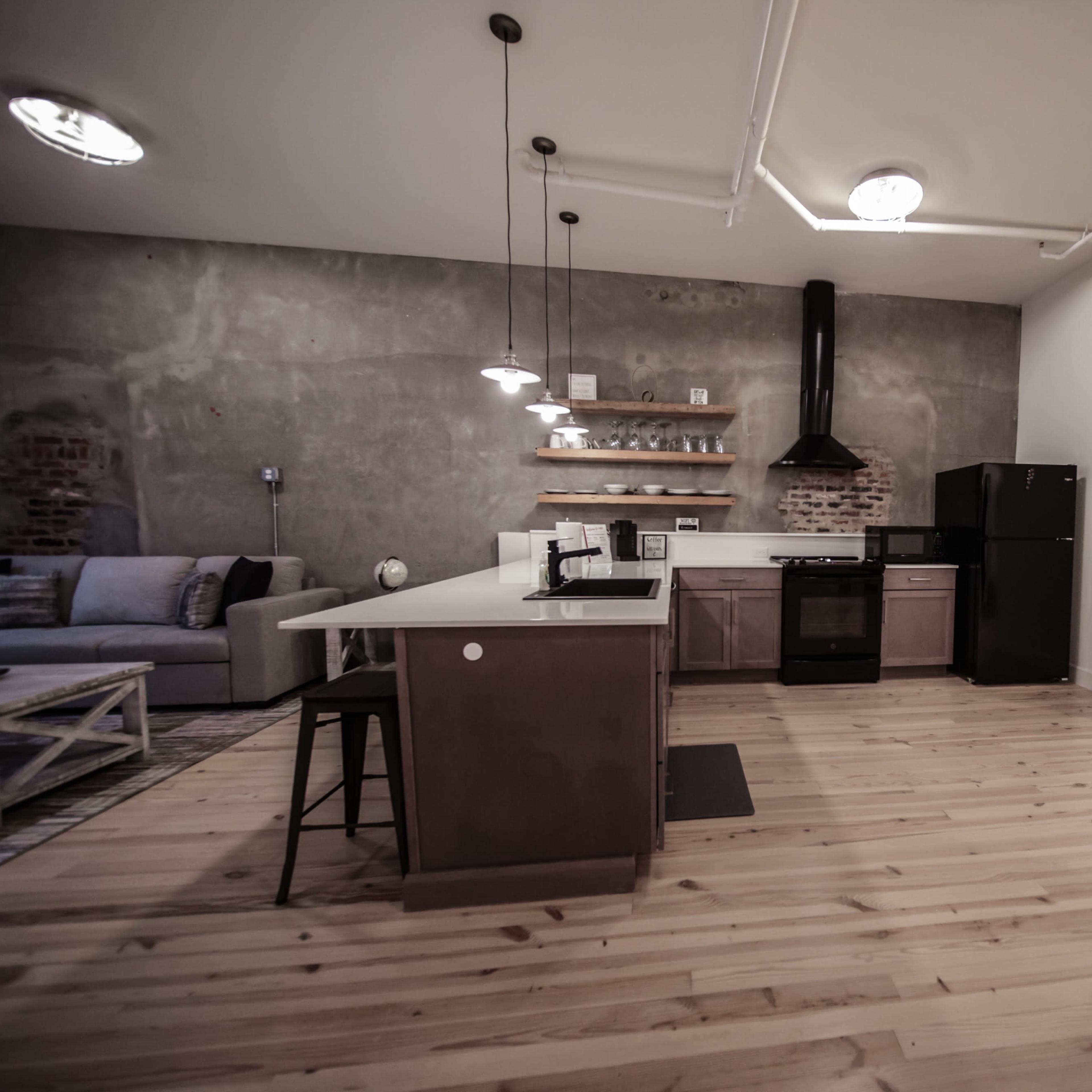 A modern kitchen and living area with concrete walls, a black stove, a refrigerator, and a wooden dining table.