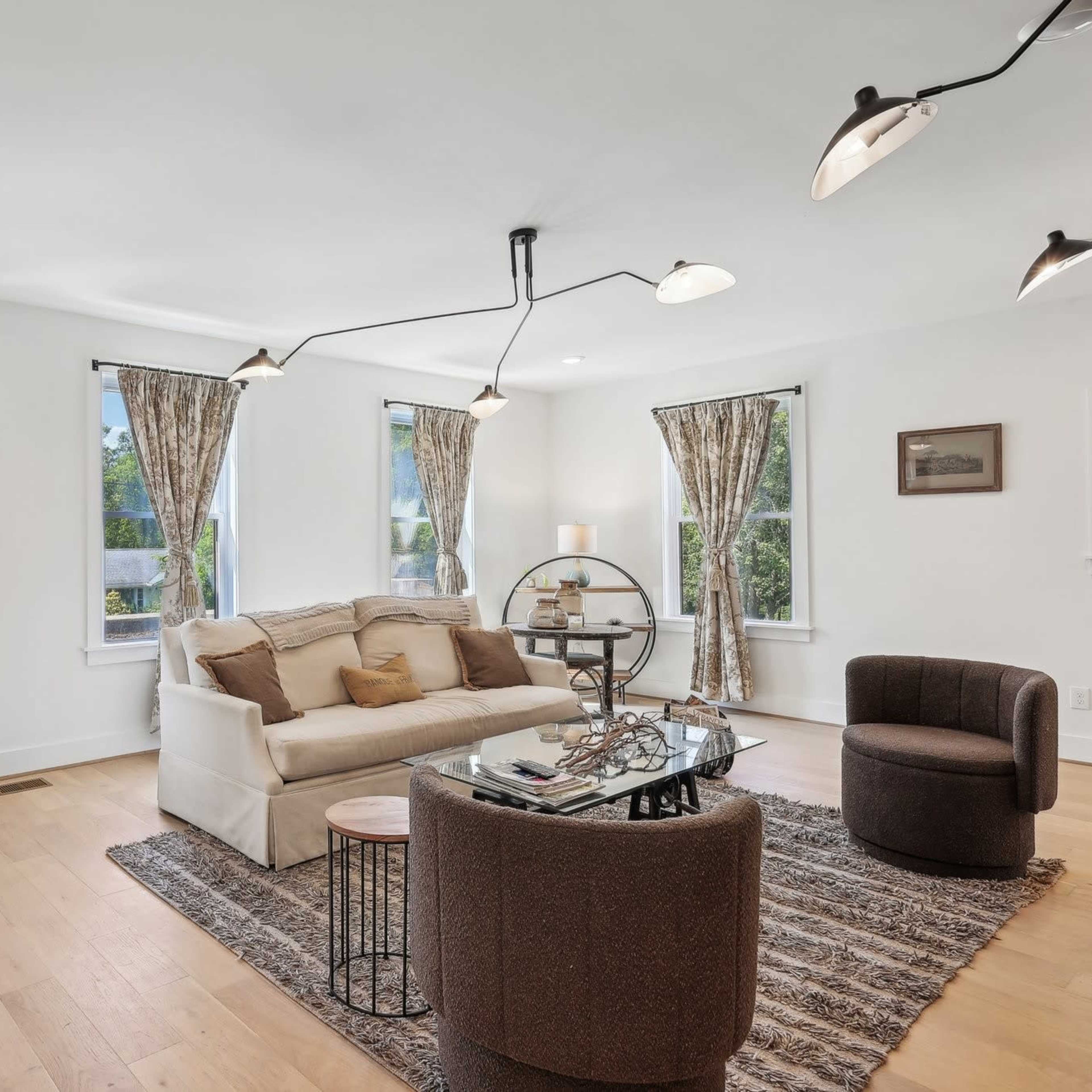 A modern living room with a white sofa, two armchairs, large windows with curtains, and a glass-top coffee table.