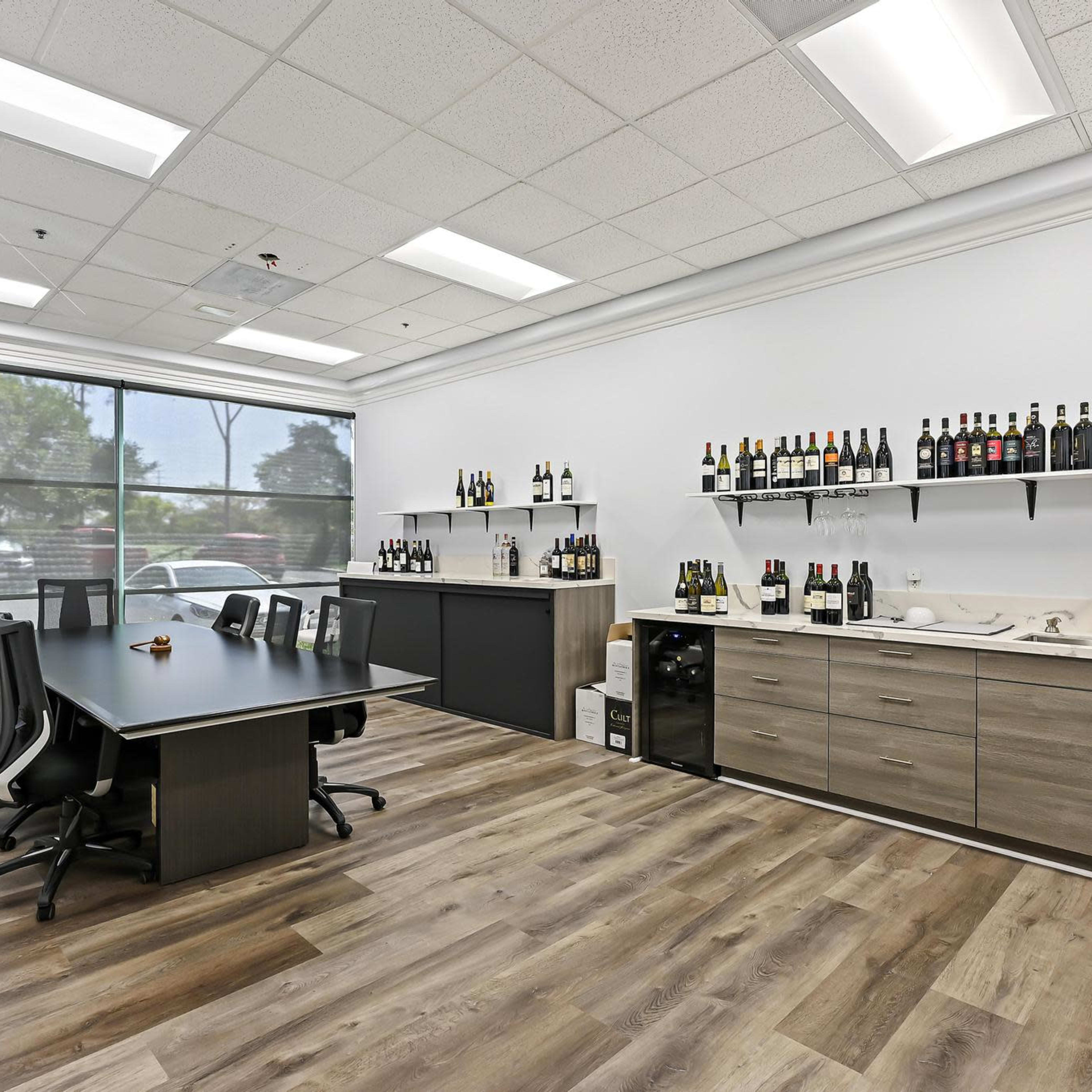A modern office space with a large table, black chairs, a counter displaying various bottles, and a large window providing natural light.