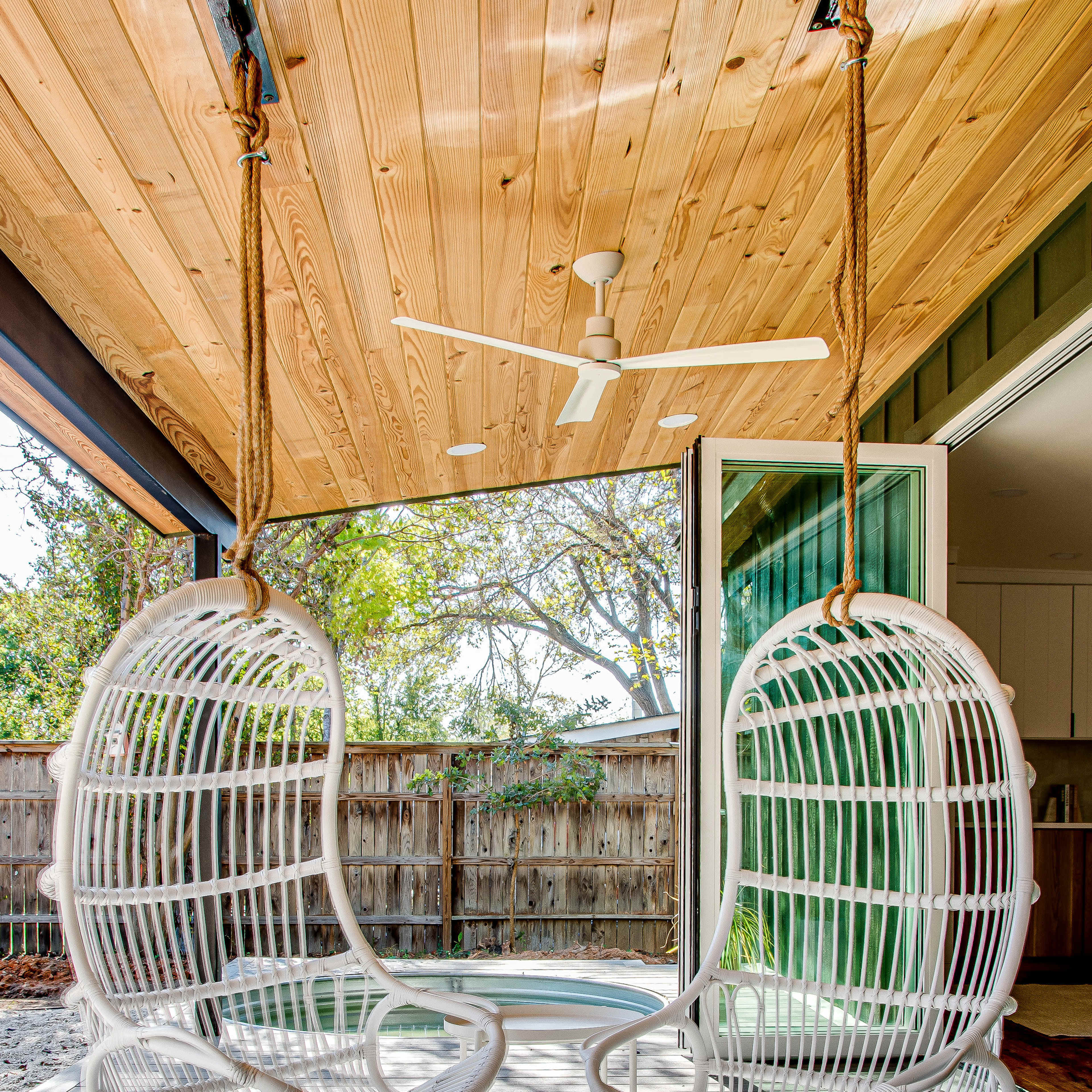 Two white hanging chairs are suspended from a wooden ceiling in a bright, open space with a view of greenery outside.
