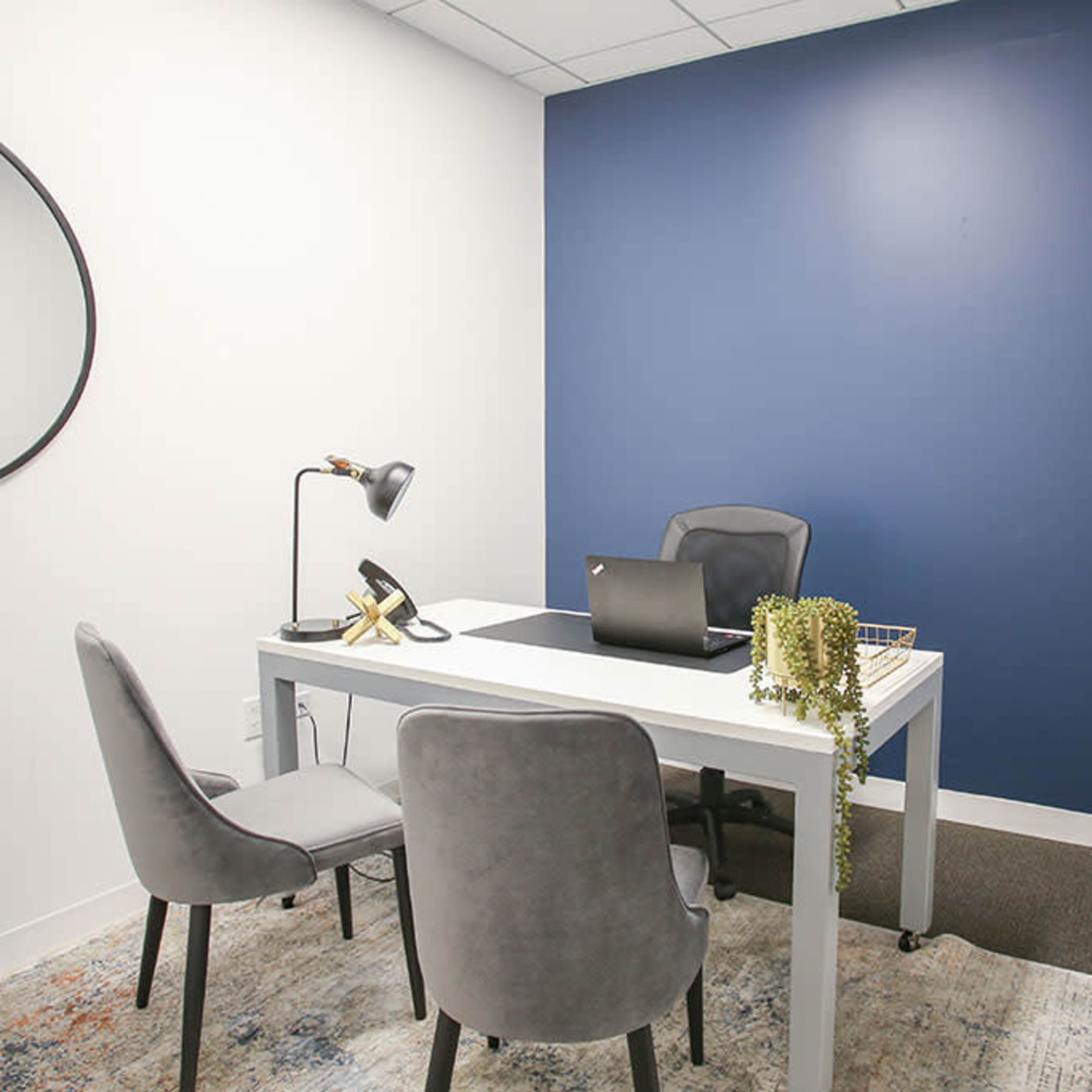 A small office with a white wall on one side and a dark blue wall on the other, featuring a desk, two gray chairs, a lamp, and a laptop.