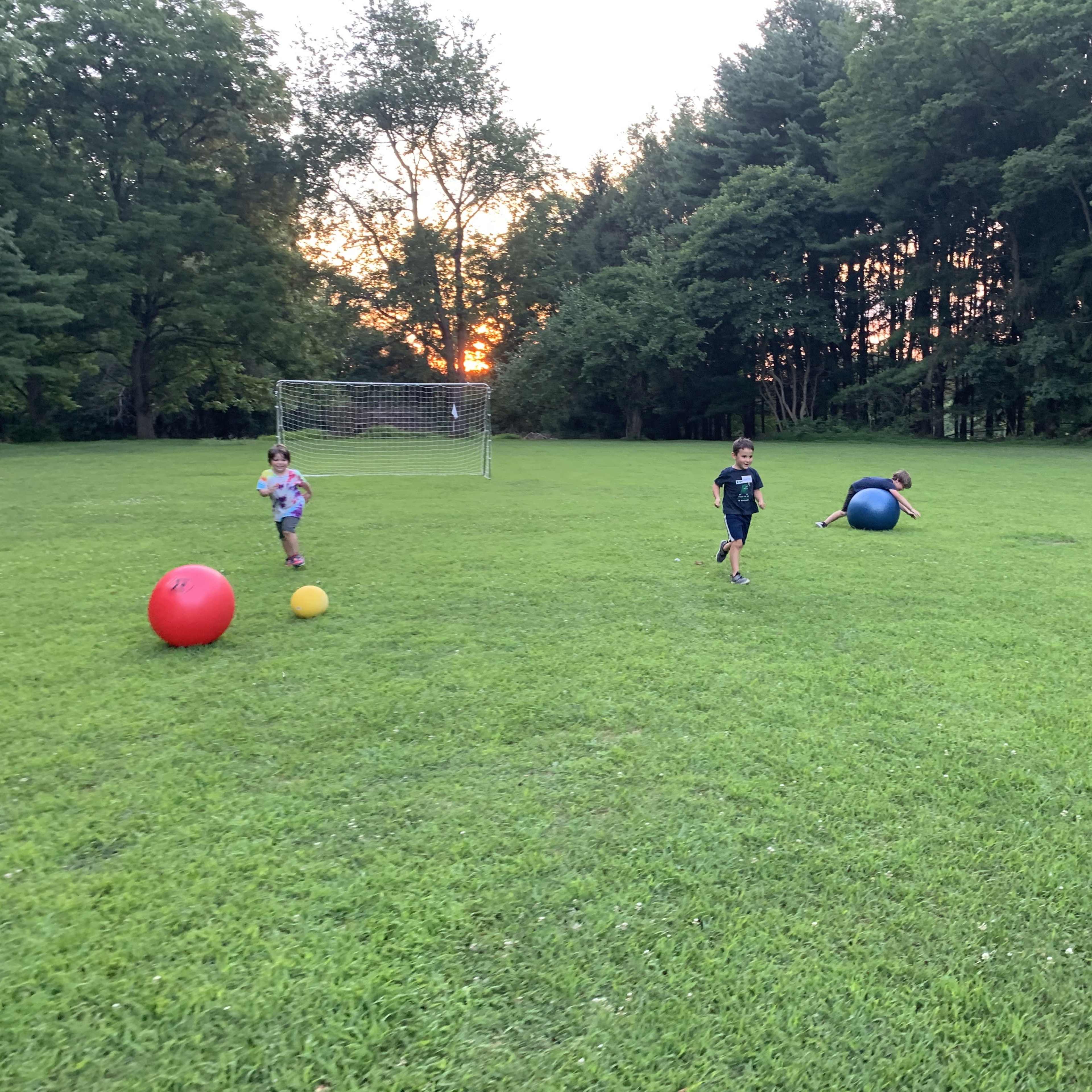 Three children are playing with colorful balls on a grassy field during sunset.