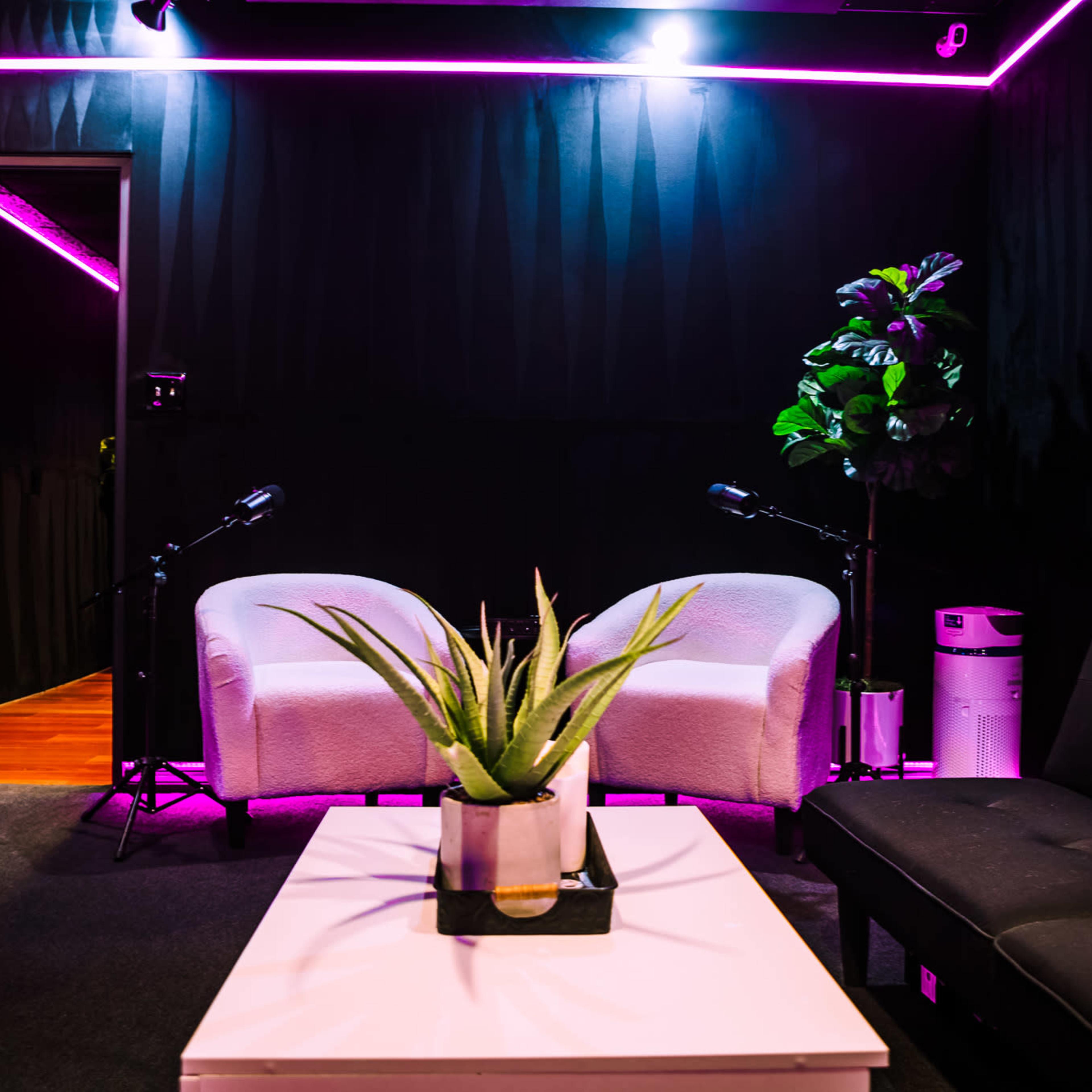 A cozy room features two white chairs, a black futon, a small plant on a coffee table, and a microphone setup under pink neon lighting.
