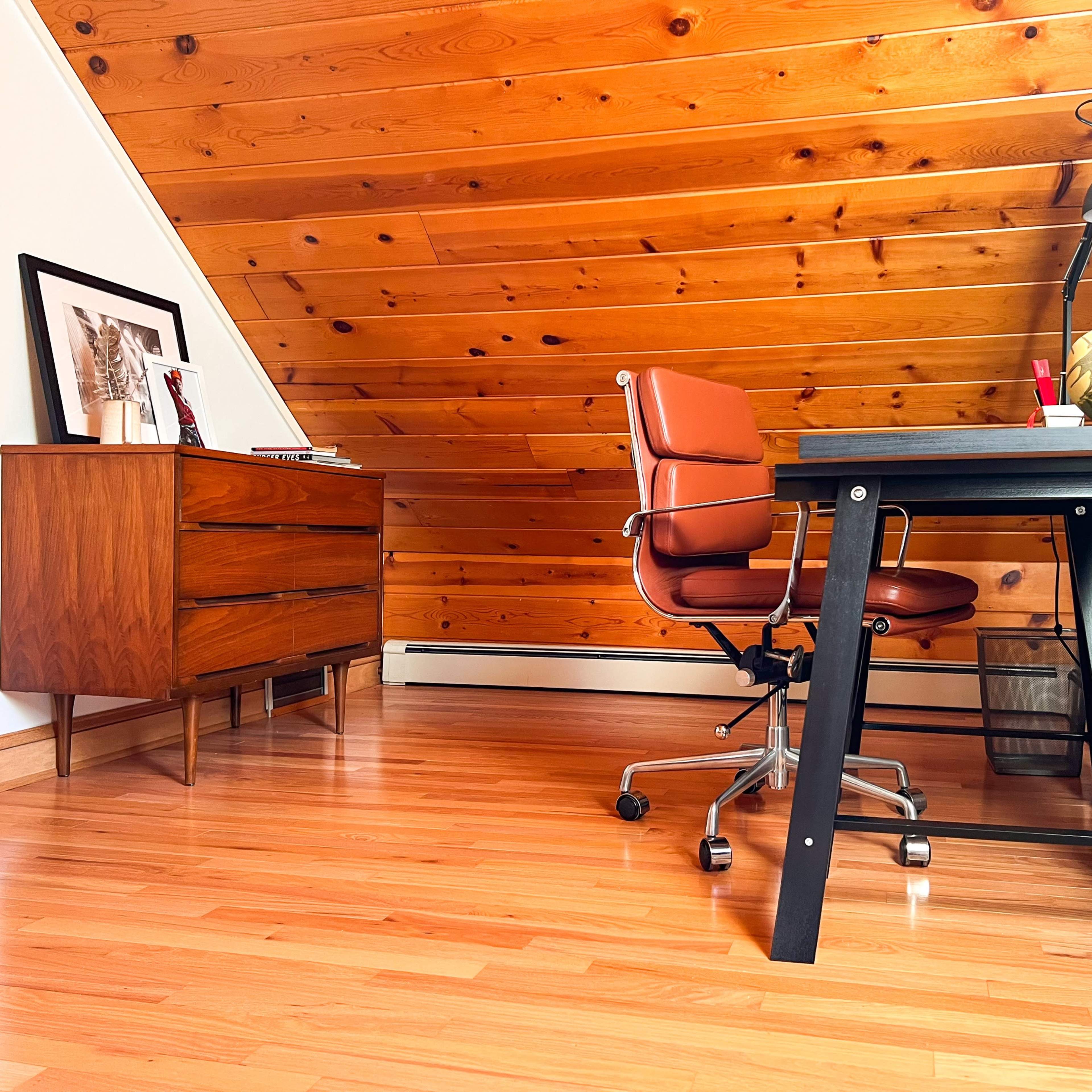 A minimalist home office features a wooden desk and an ergonomic chair, with a vintage dresser and sloped wooden ceiling in the background.