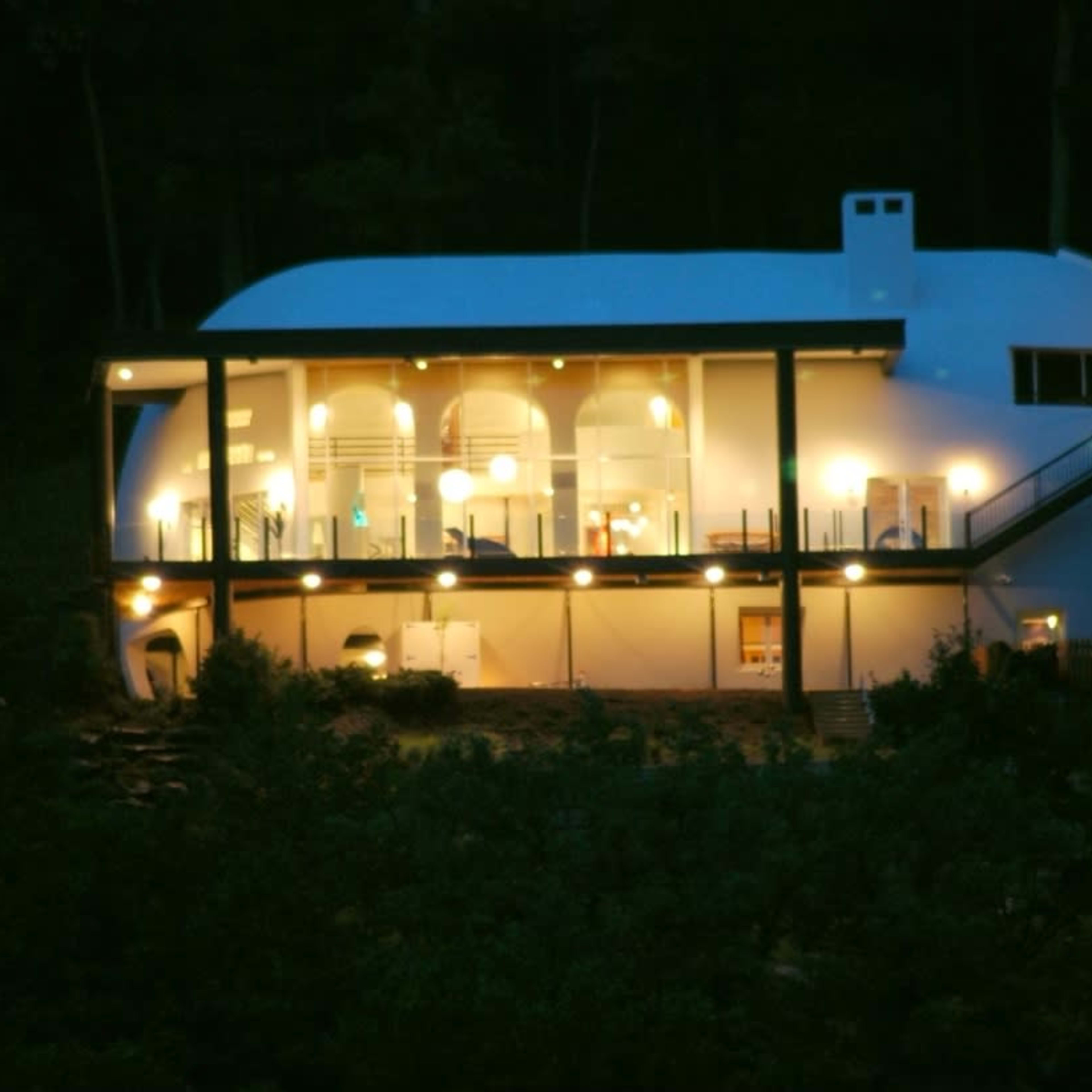 A modern, white, rounded house is illuminated at night, featuring multiple balconies and large windows overlooking a wooded area.