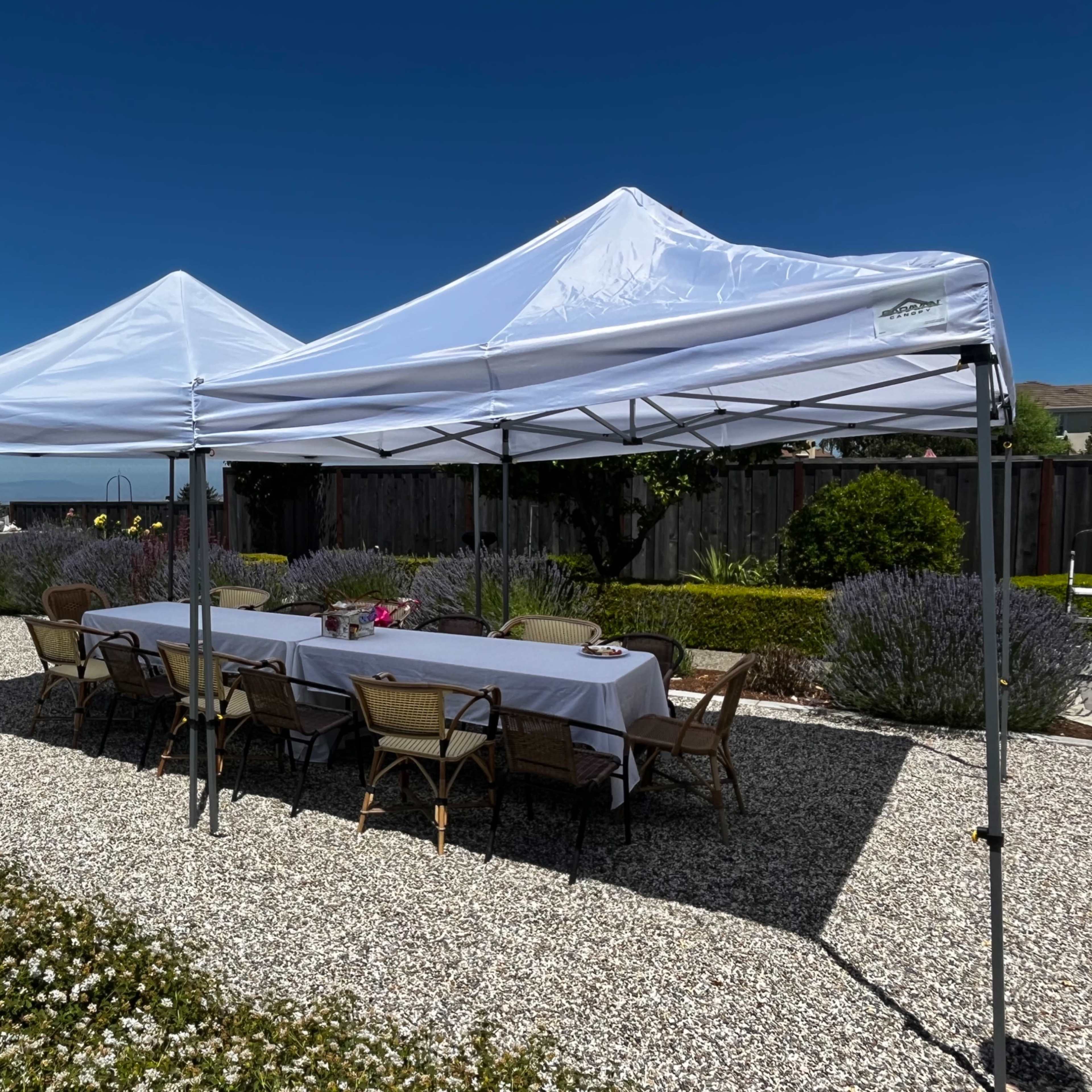 Two white canopies are set up over a long table surrounded by chairs on a gravel patio with landscaping in the background.