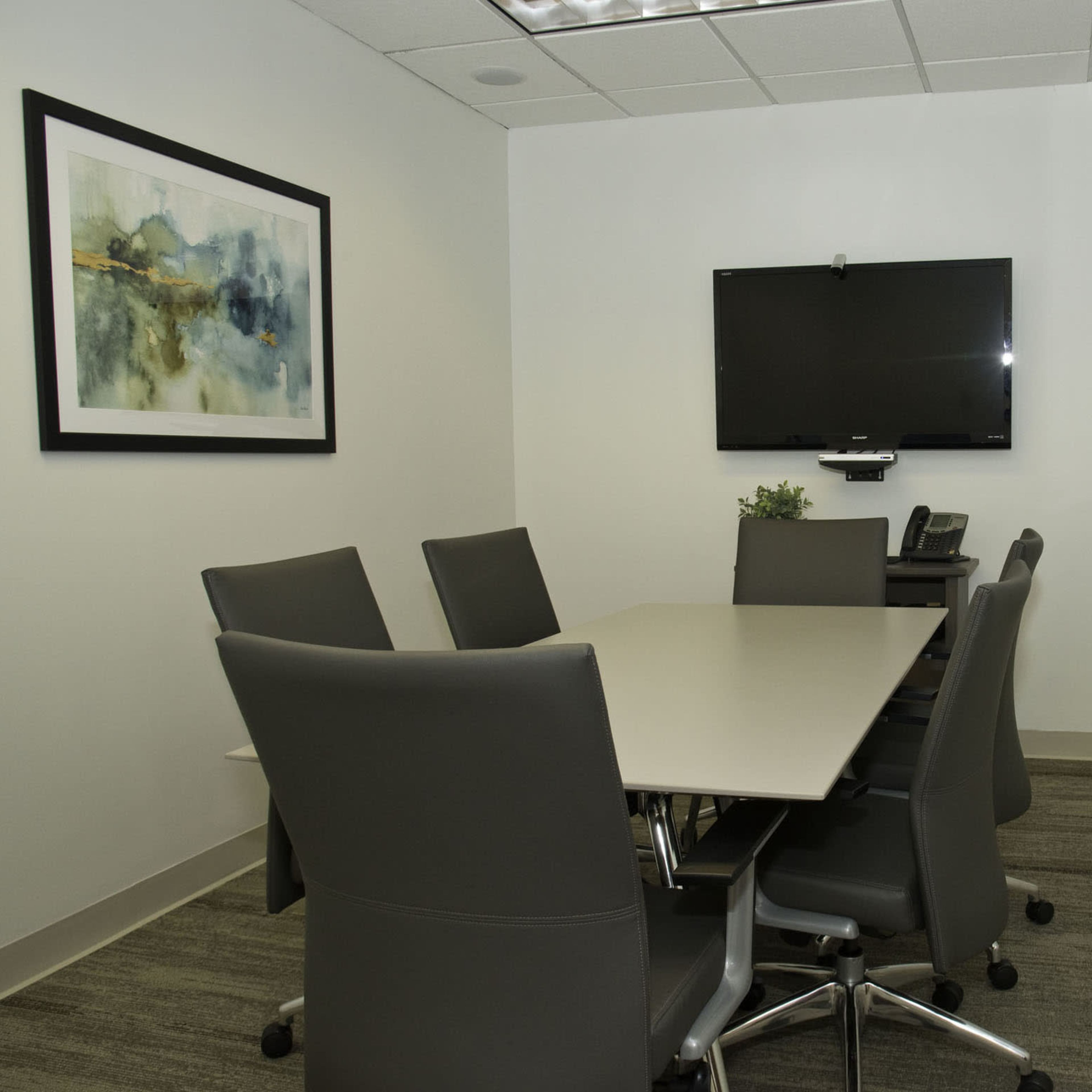 A small conference room with a rectangular table surrounded by six chairs, a wall-mounted television, and a framed artwork.