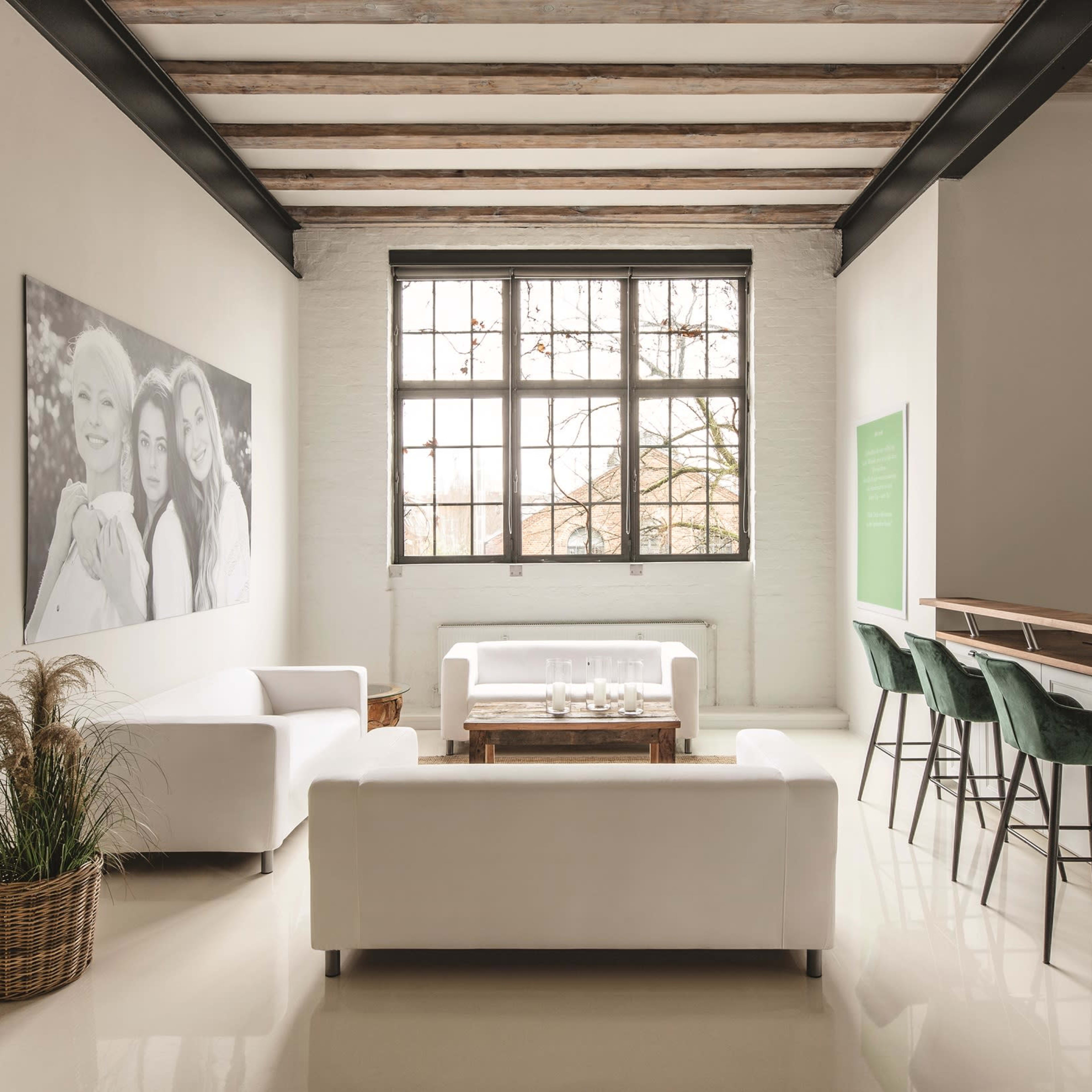 A minimalist living space with two white sofas, a wooden coffee table, bar seating with green stools, and large windows allowing natural light.