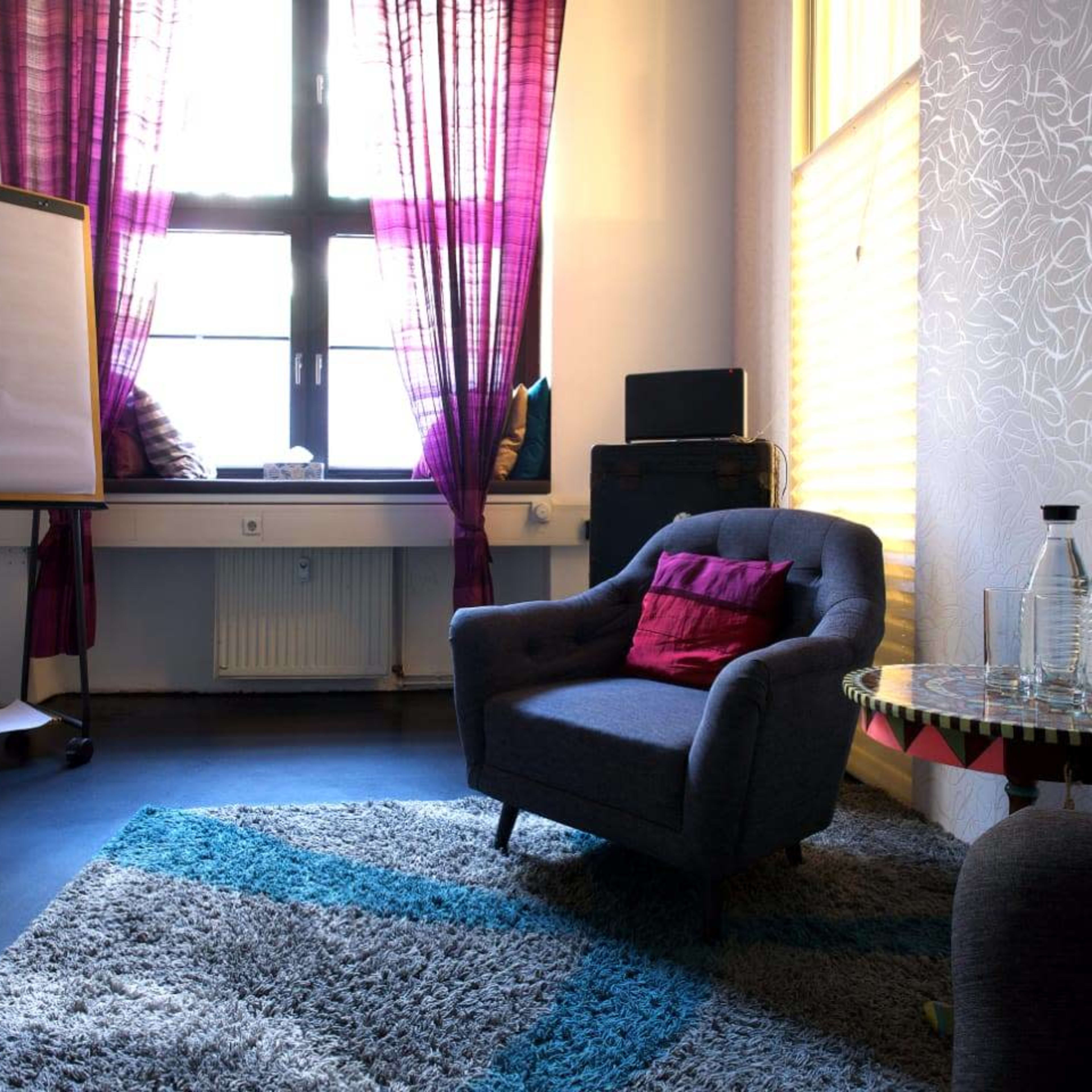 A cozy room features a gray armchair, a table with glass bottles, a flip chart stand, and vibrant curtains by a large window.