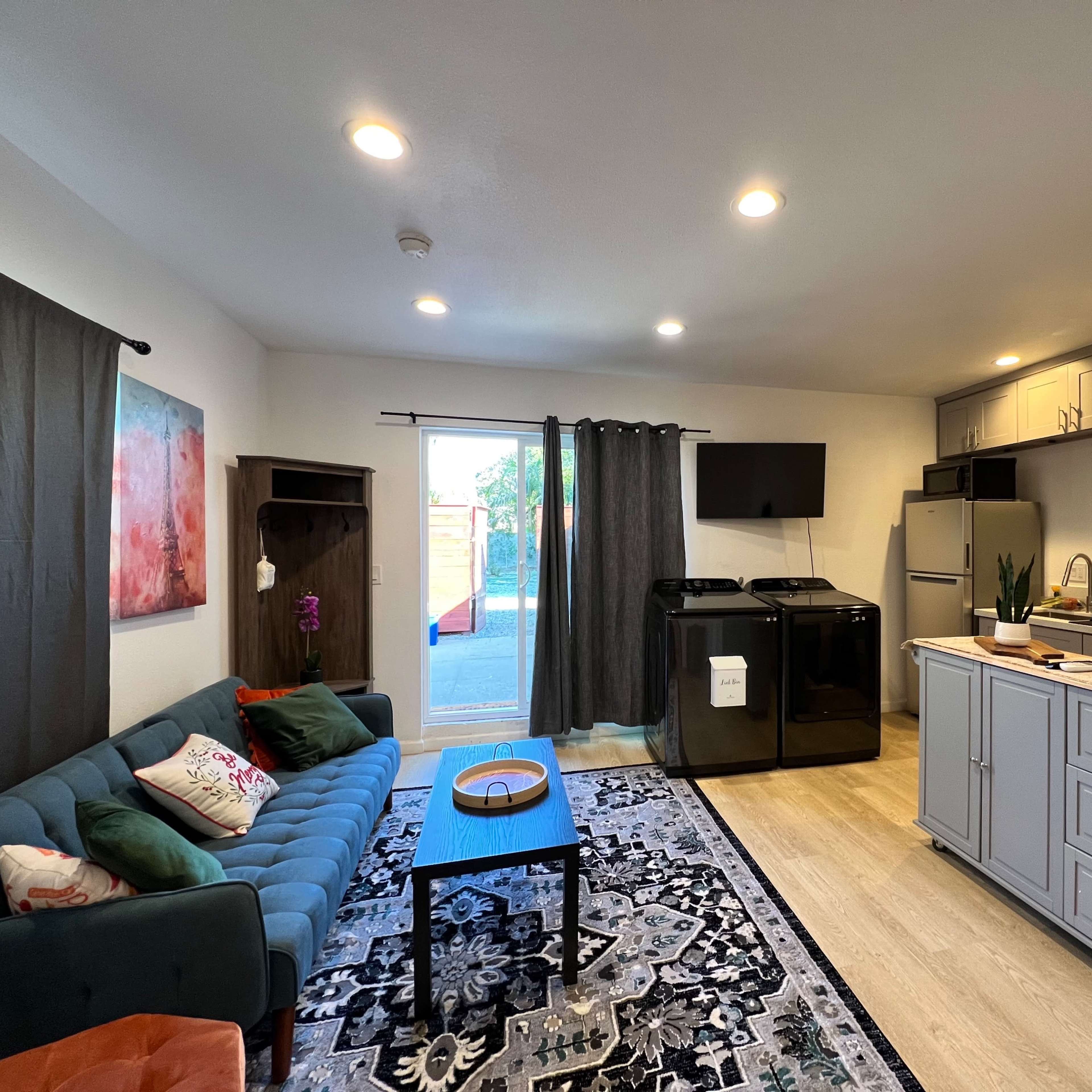 A modern open-concept living space featuring a kitchen with gray cabinets, a black refrigerator, and a comfortable seating area with a teal couch and decorative rug.