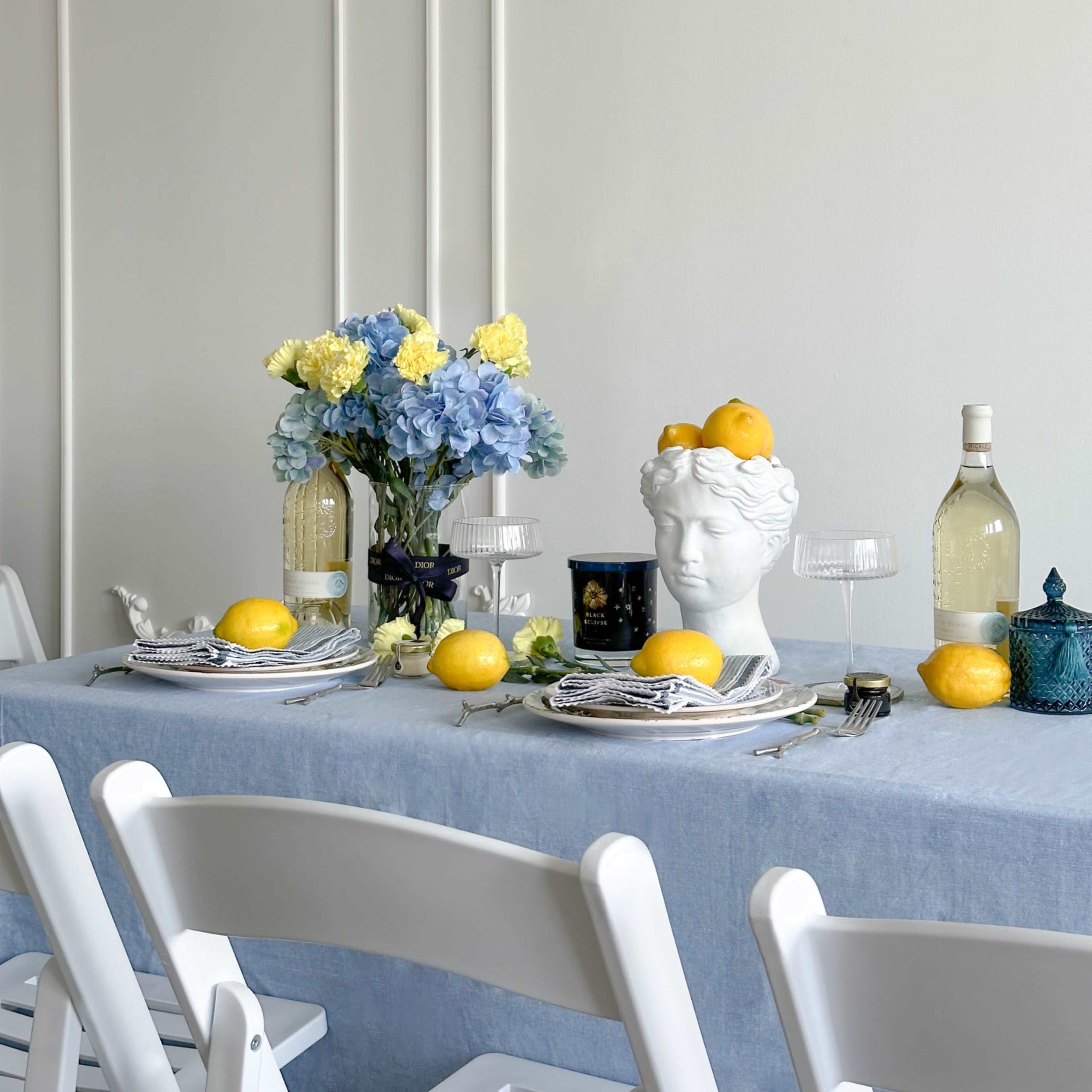 A dining table is set with a blue tablecloth, decorative flowers, and a white bust centerpiece adorned with lemons, flanked by wine bottles and glasses.