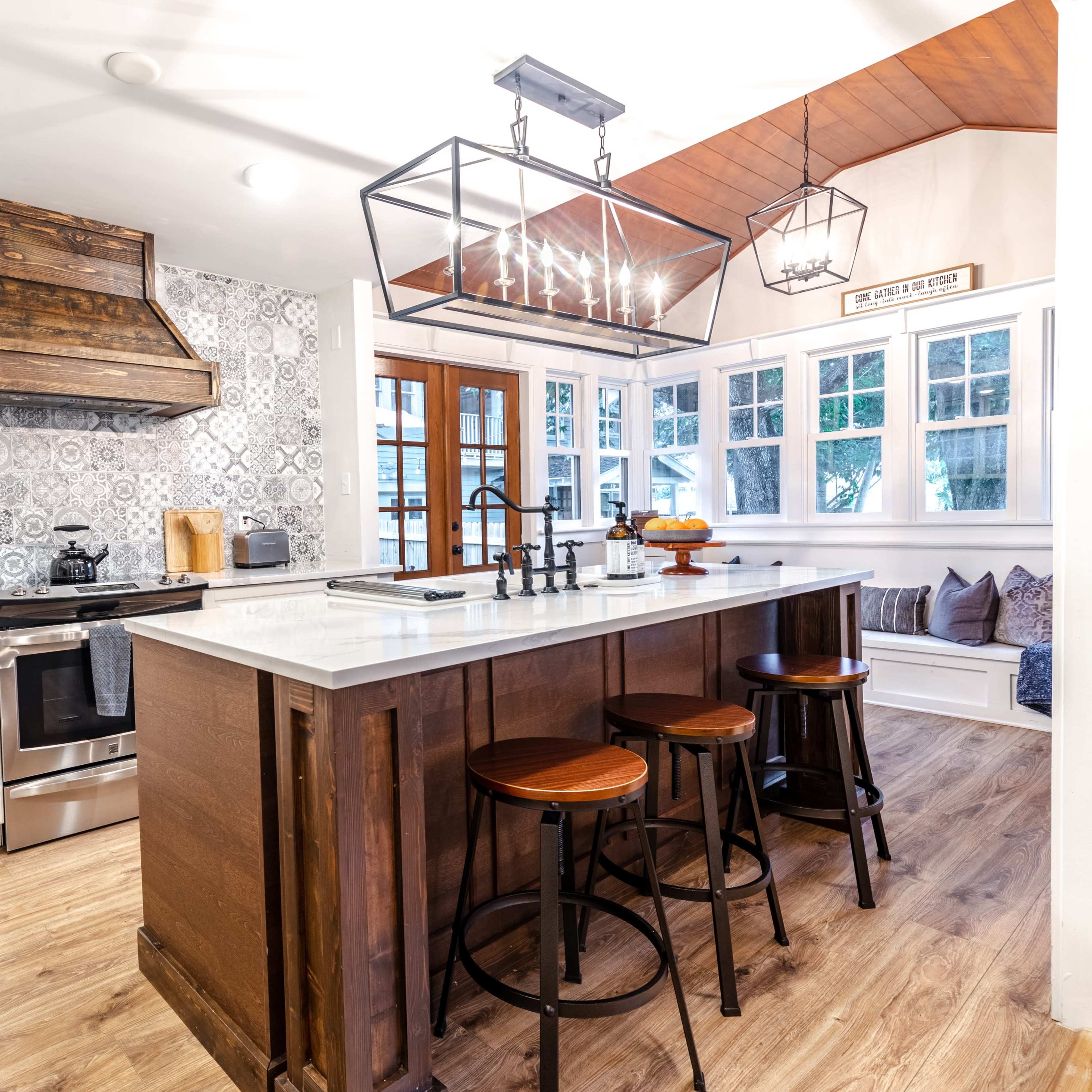 A modern kitchen featuring a central island with three stools, a stainless steel stove, and patterned tile backsplash.