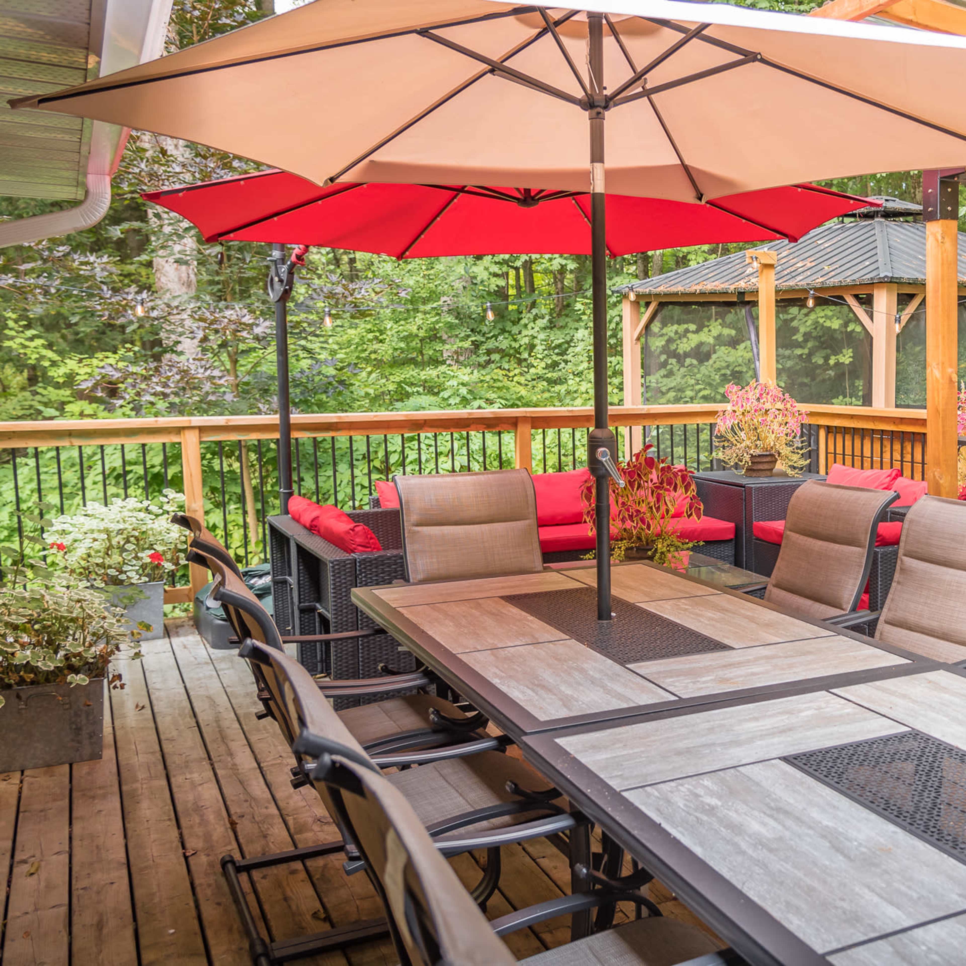 A wooden deck features a dining table with multiple chairs, an umbrella for shade, and a barbecue grill, surrounded by potted plants and a gazebo in the background.