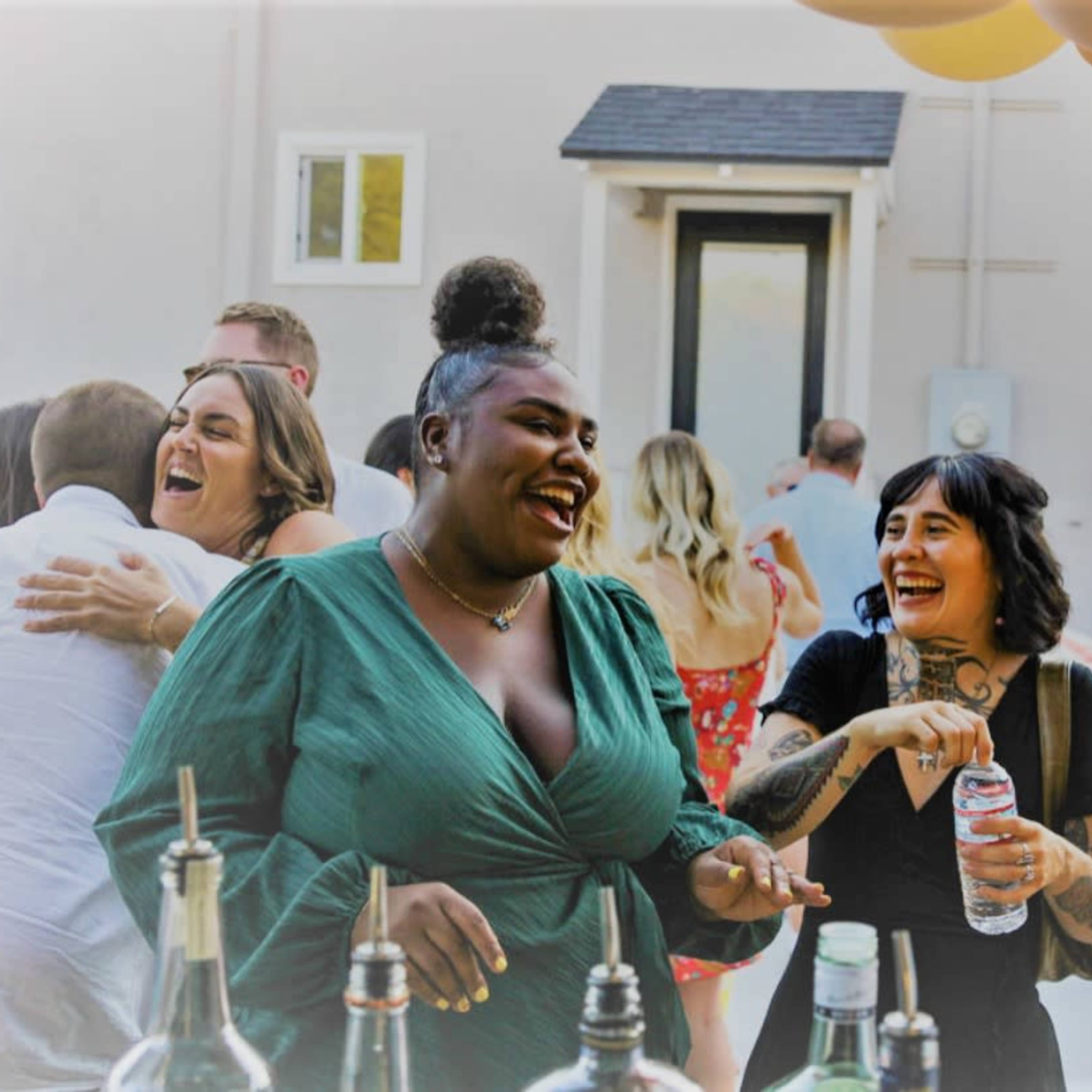 A group of friends enjoys drinks and laughter at a festive outdoor gathering near a bar setup with balloons in the background.