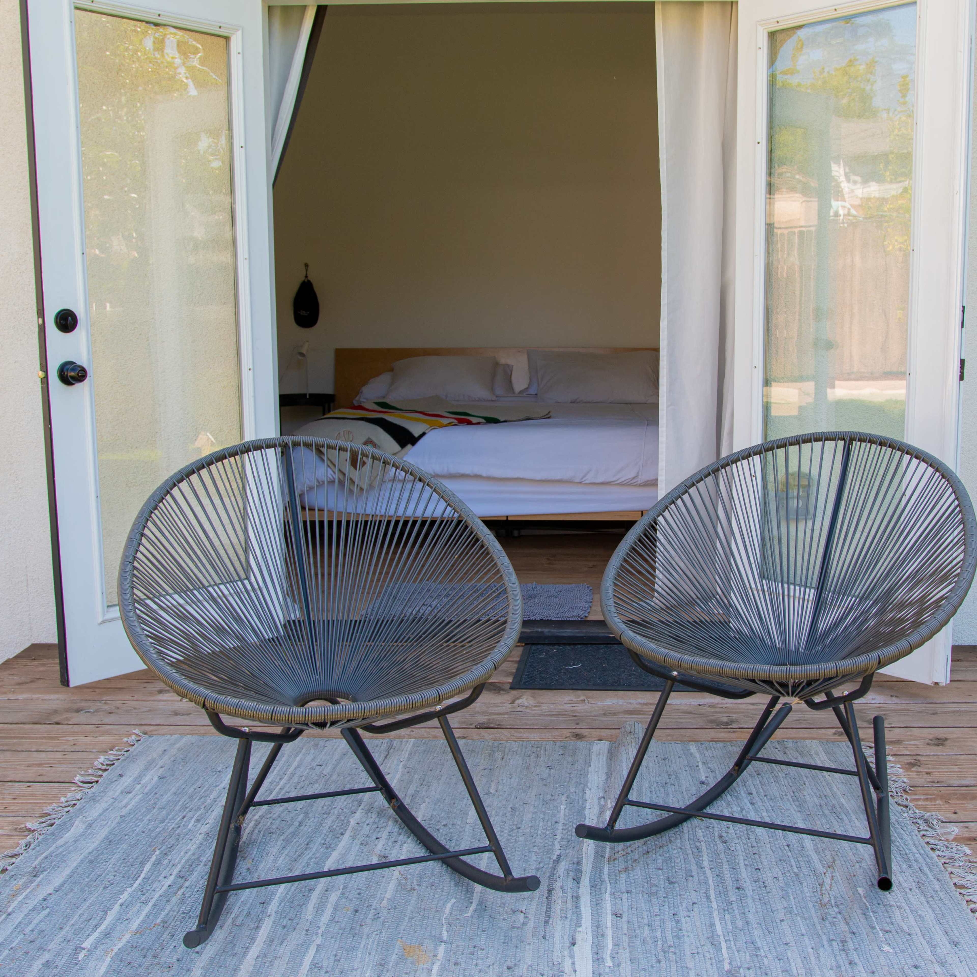 Two modern chairs are positioned on a wooden deck in front of an open doorway leading into a minimalist bedroom.