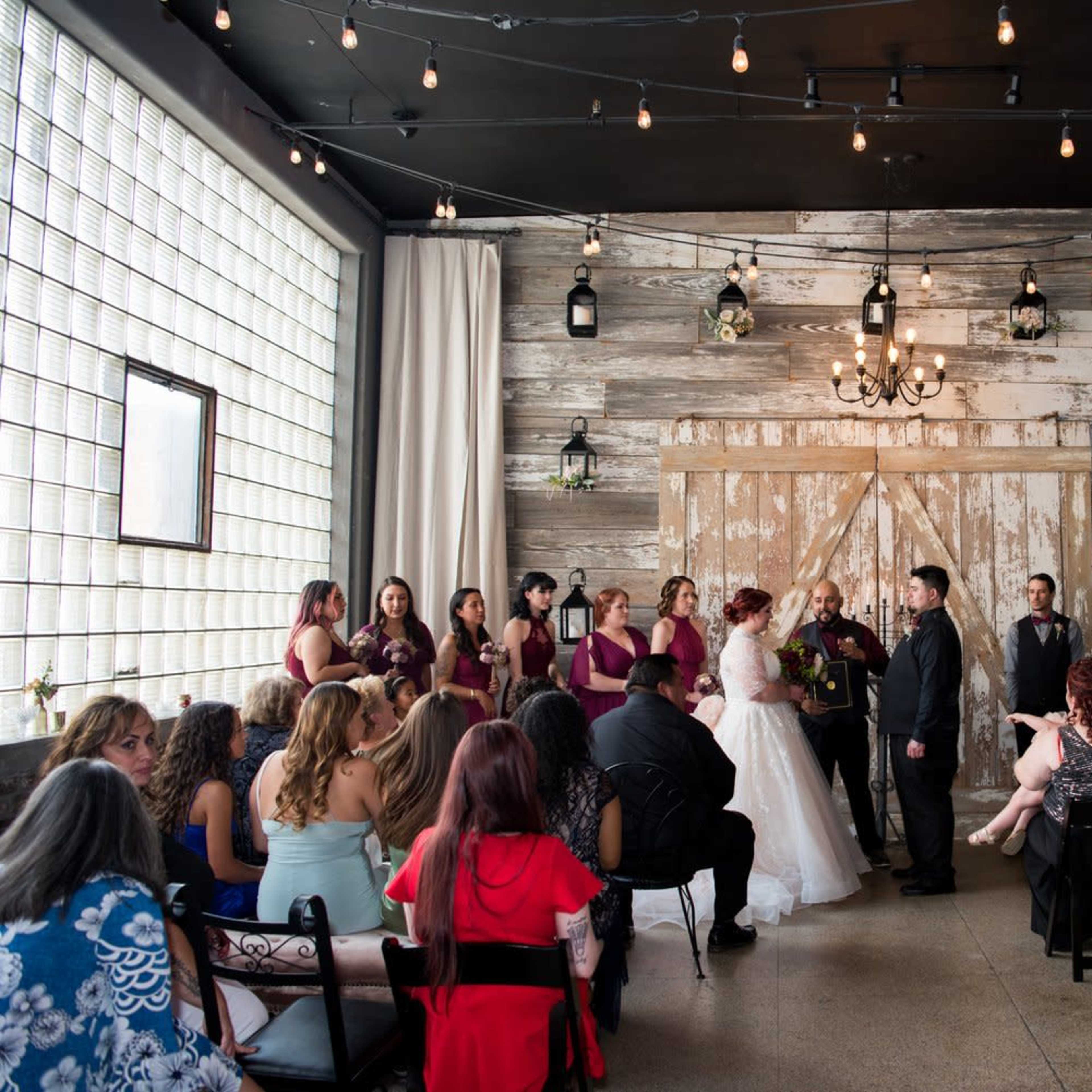 A wedding ceremony takes place indoors, with a couple standing at an altar and guests seated nearby, surrounded by rustic decor and string lights.
