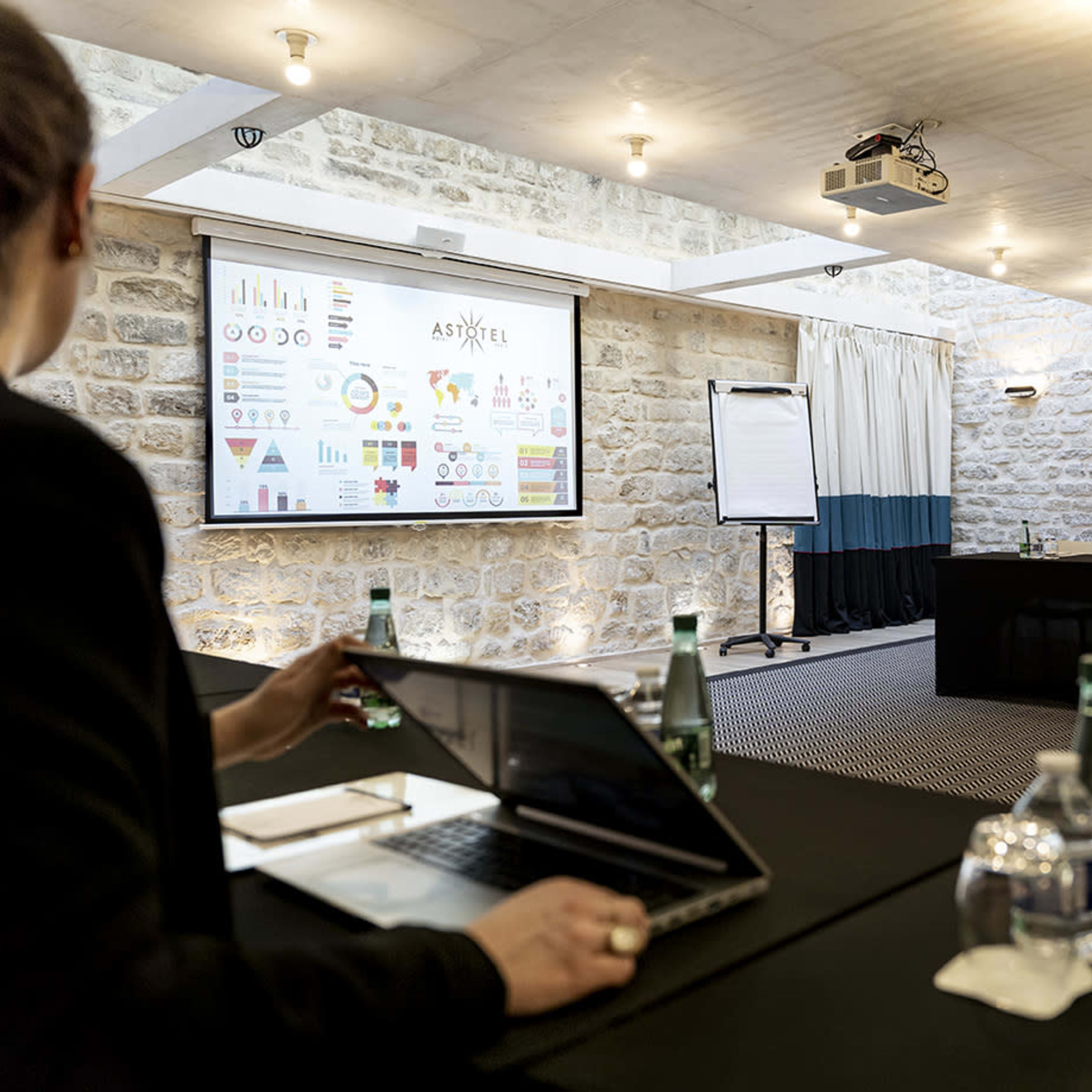 A person sits at a table in a conference room, viewing a presentation displayed on a screen, with multiple water bottles and laptops on the table.