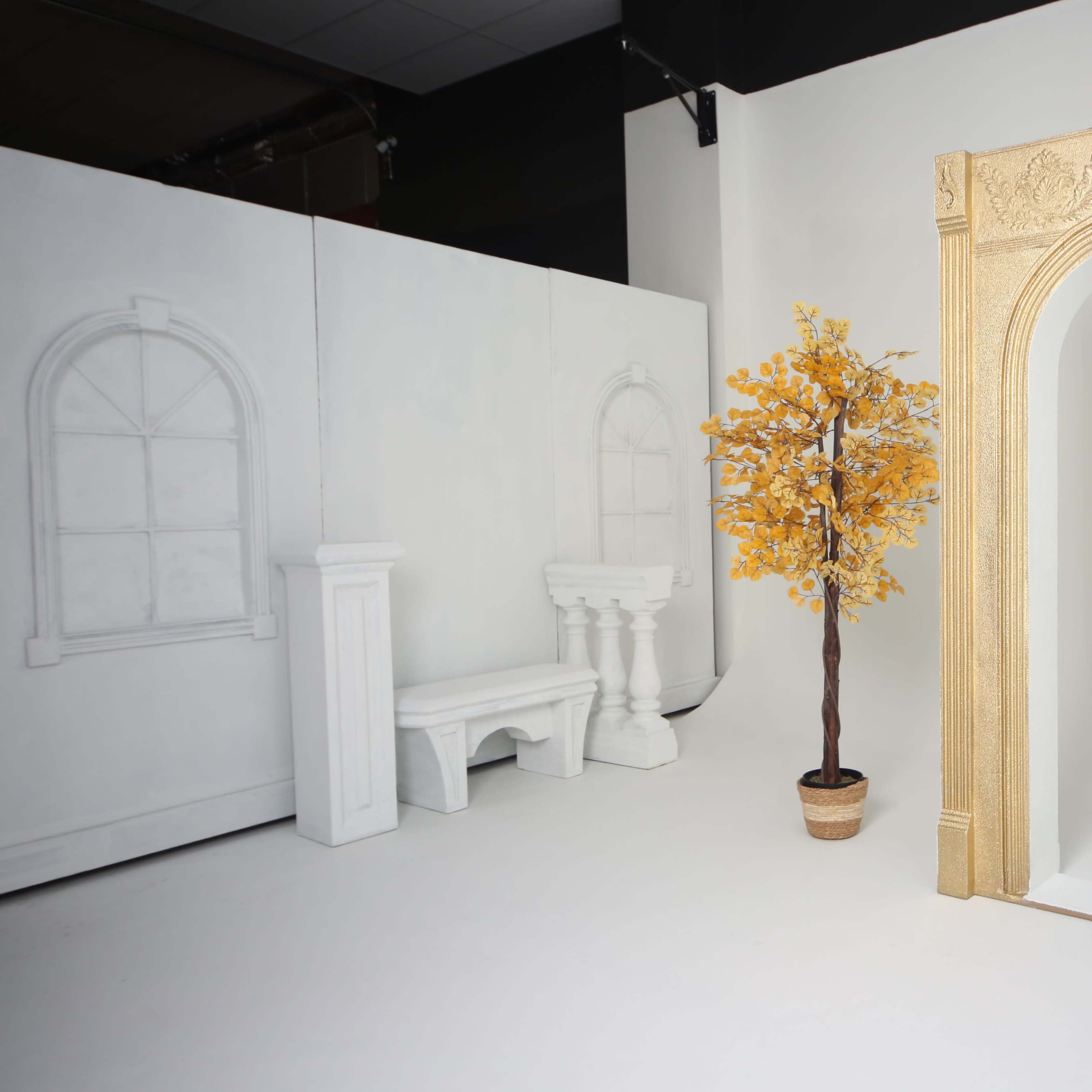 A stylized gold archway next to a small potted tree with yellow leaves, positioned in a minimalistic white room with a partial wall featuring a window outline.