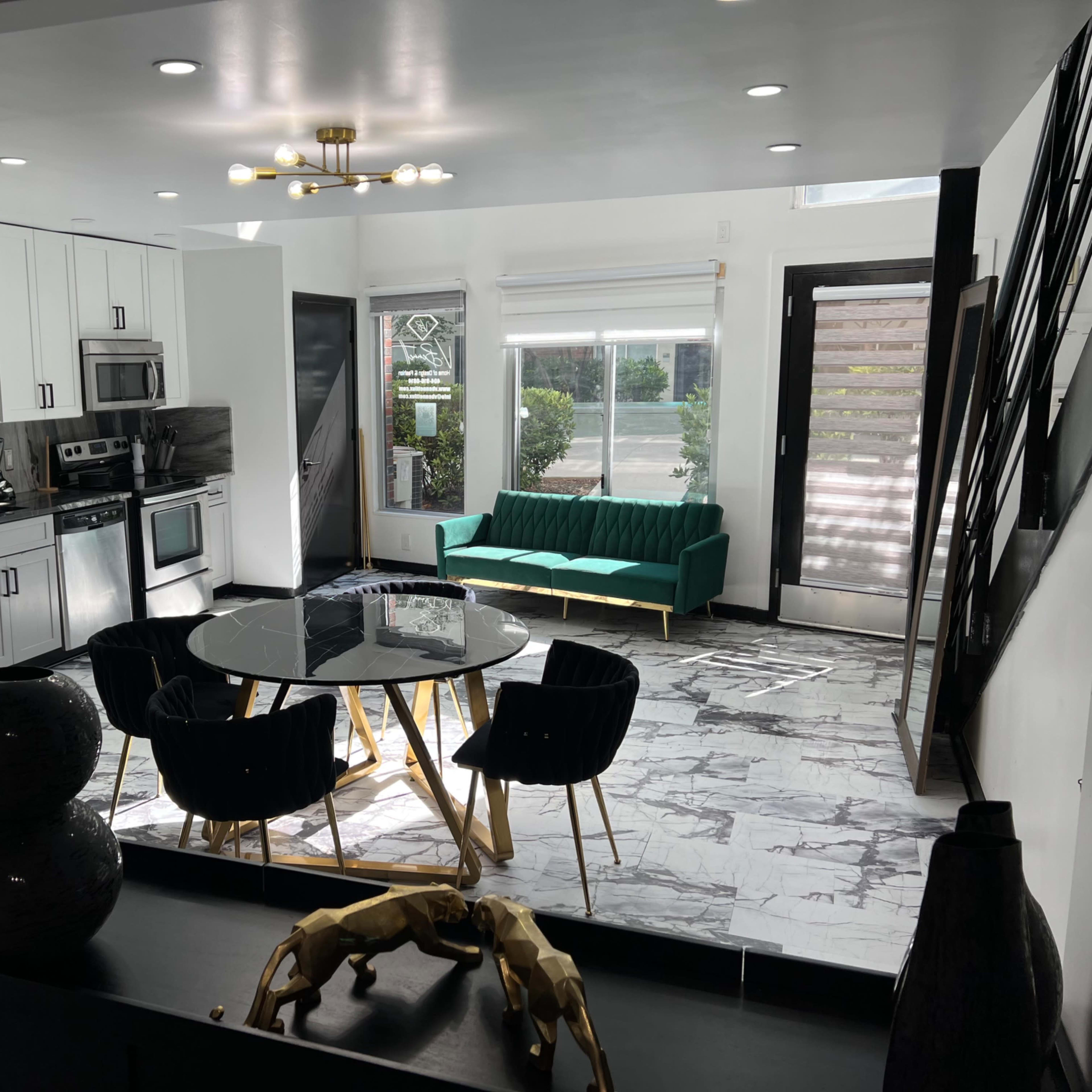 A modern kitchen and living area featuring a black and white marble floor, a green couch, a round dining table with black chairs, and stainless steel appliances.