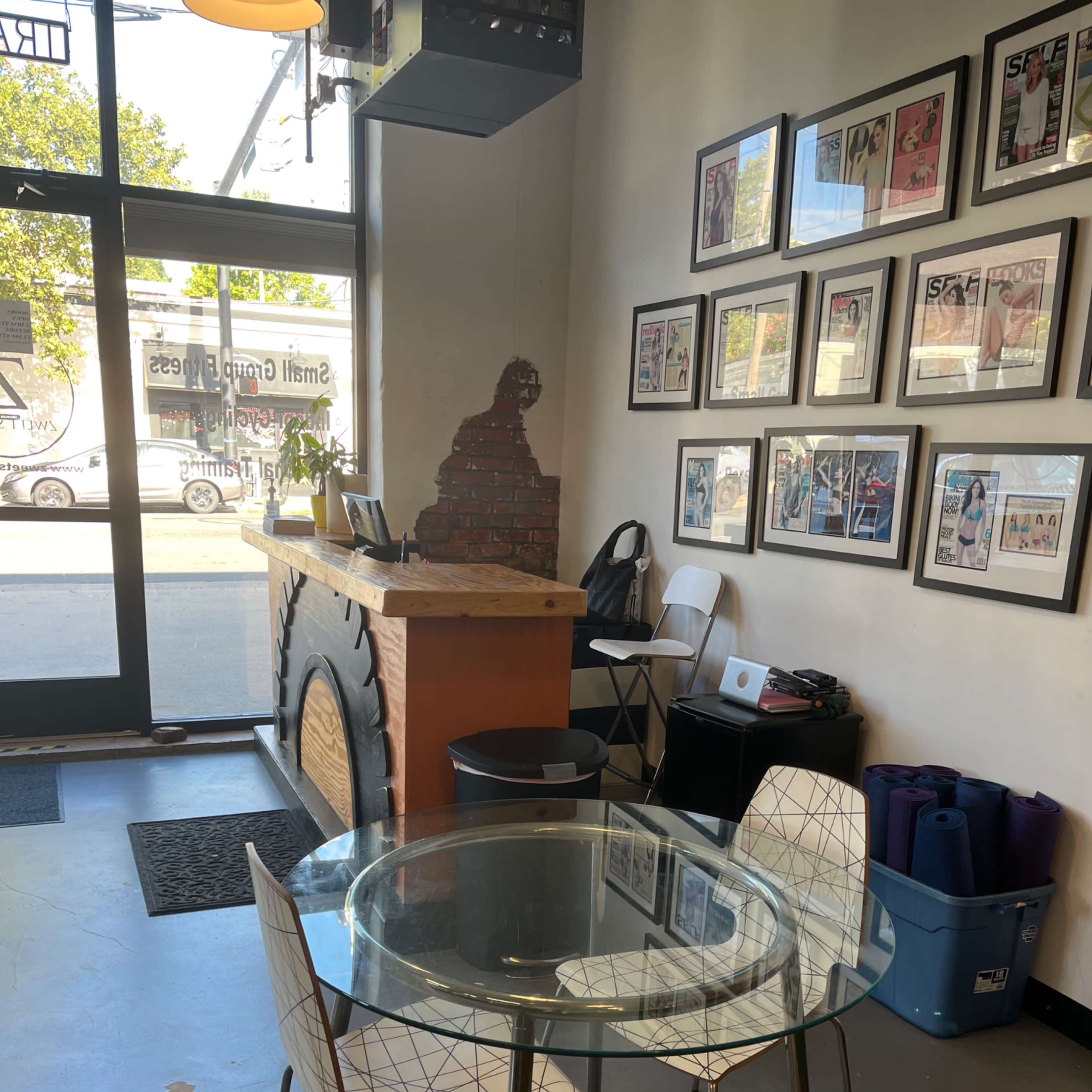 A small interior space with a glass table and two chairs, a wooden counter with magazines on the wall, and a window displaying the street outside.