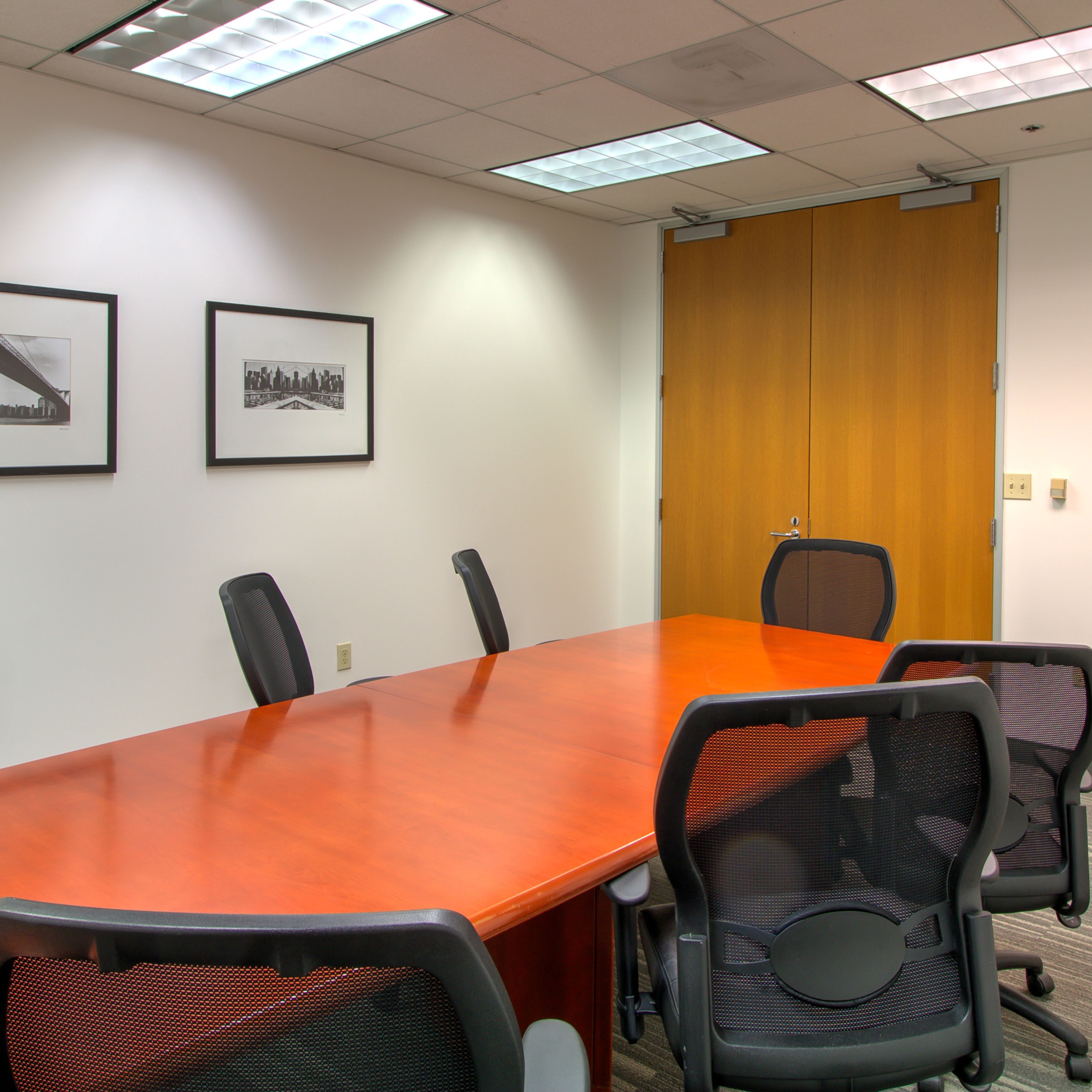 A conference room features a long wooden table surrounded by black ergonomic chairs, with framed black-and-white photographs on the walls and a clock above a side table.