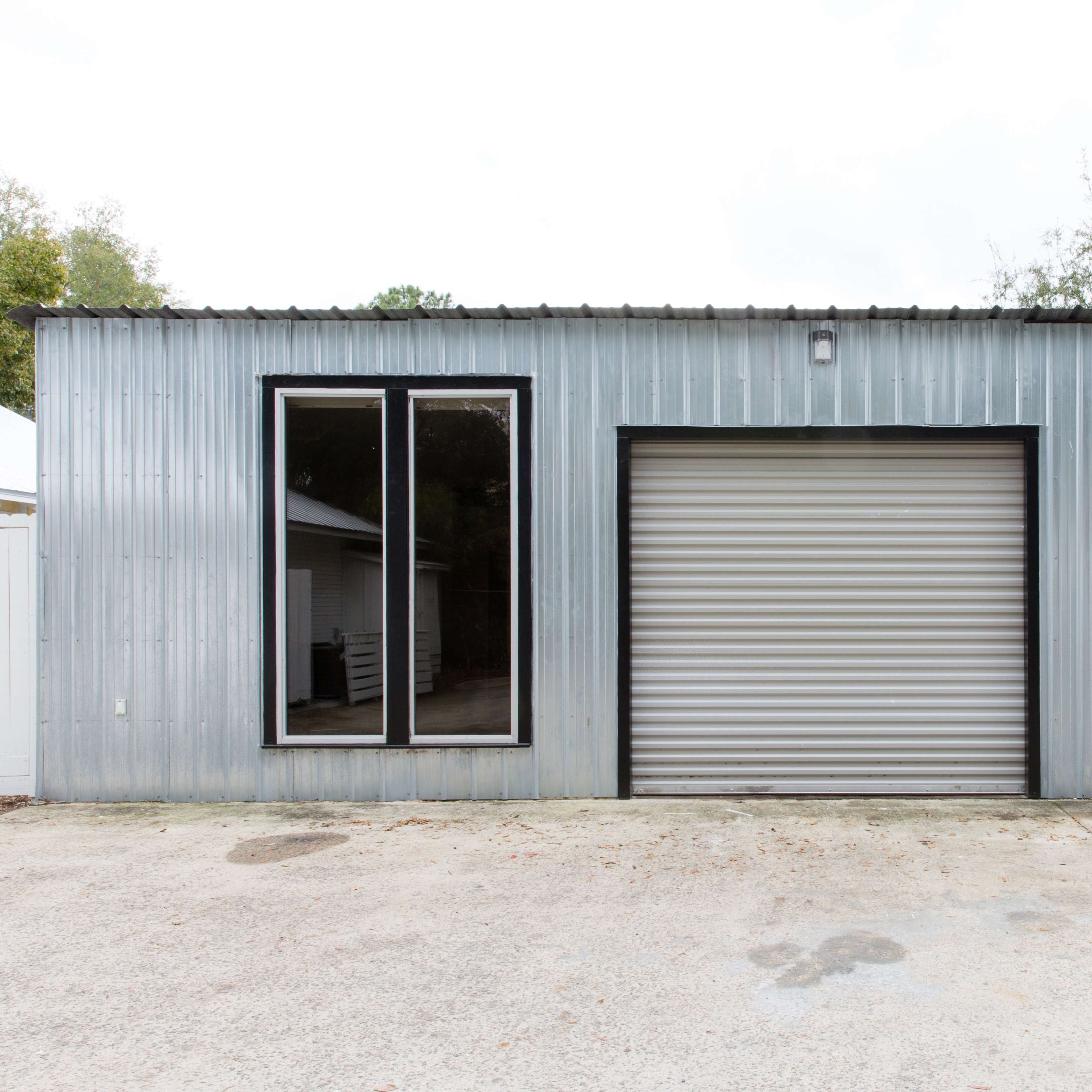 A metallic, industrial-style building with a large roll-up door, a single black door, and a large window on one side.