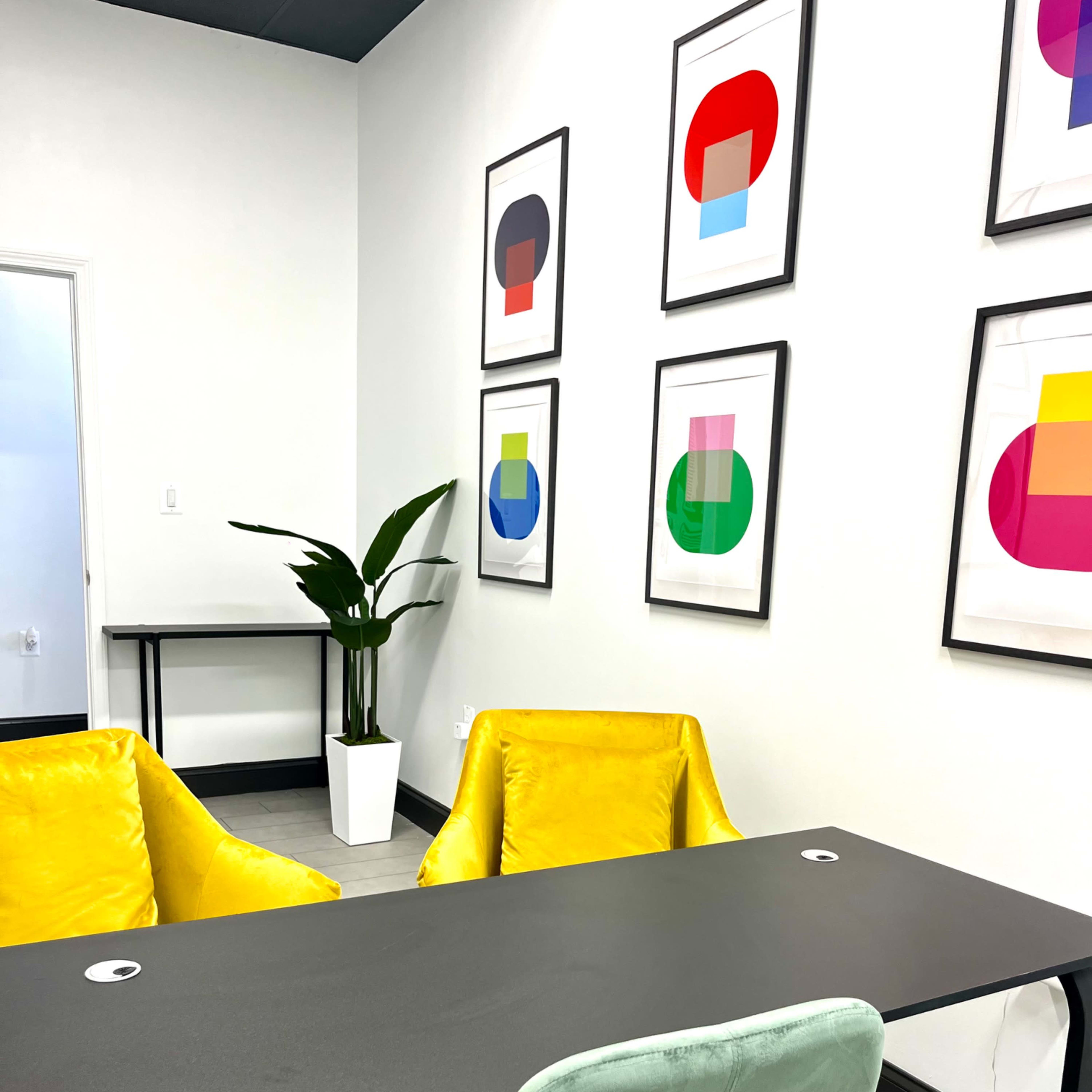 A modern office with two yellow chairs and a green office chair facing a black desk, while colorful abstract art pieces hang on the wall.