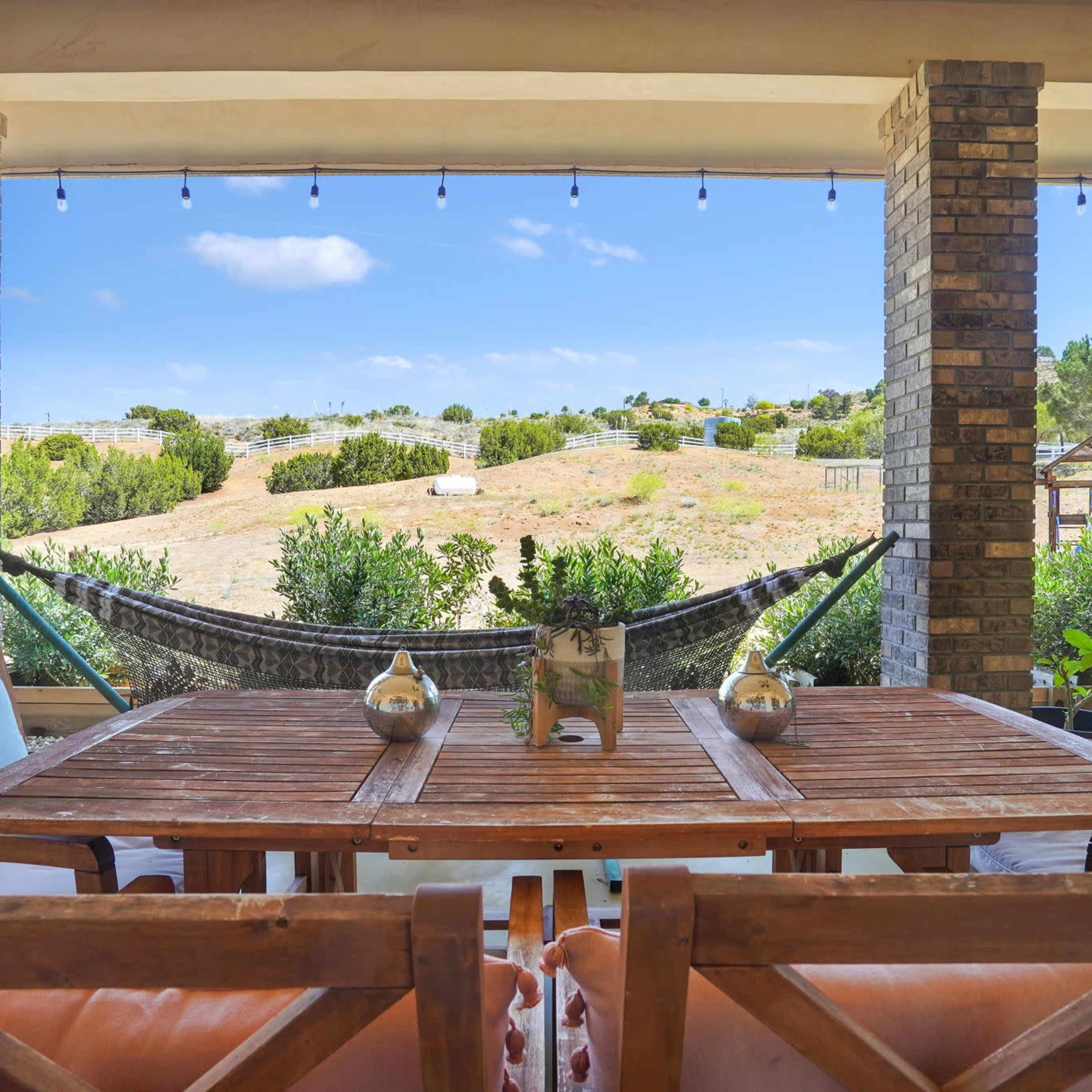 A wooden table with chairs is set under a covered porch, overlooking a grassy landscape with a hammock and distant trees.
